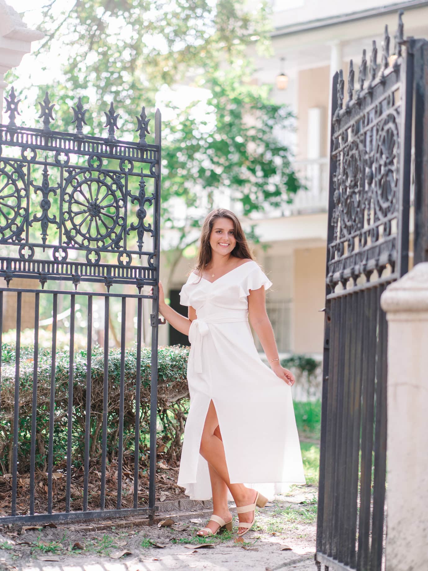 Graduating senior in white dress posing by ornate black iron gate at the College of Charleston campus