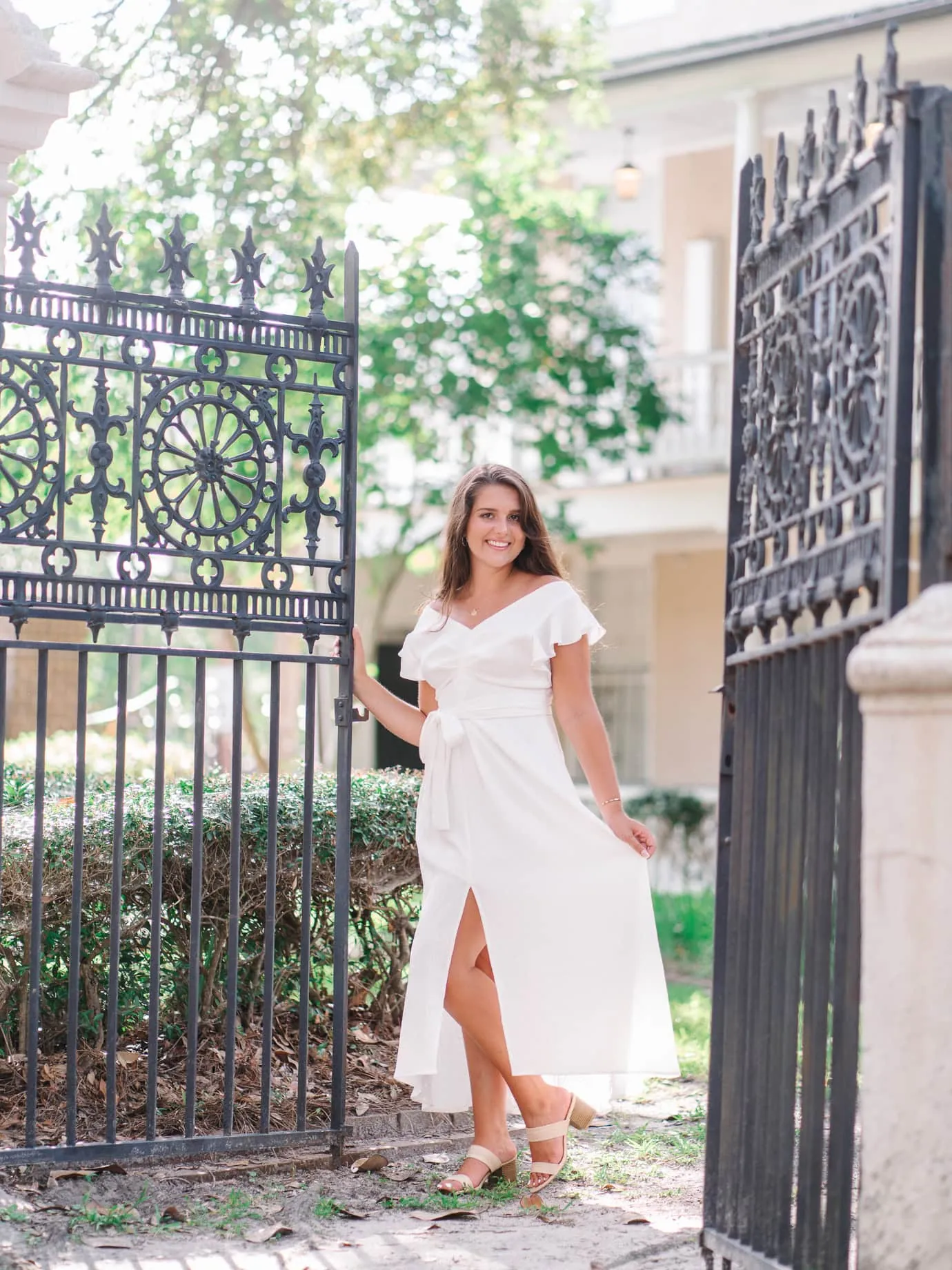 Graduating senior in white dress posing by ornate black iron gate at the College of Charleston campus