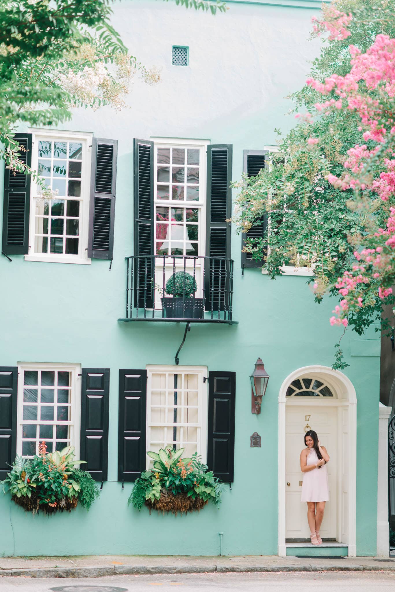 Vibrant mint green house with black shutters, pink flowering trees, and a young woman in a light pink dress standing at the door, perfect for family beach portraits and outdoor photography.