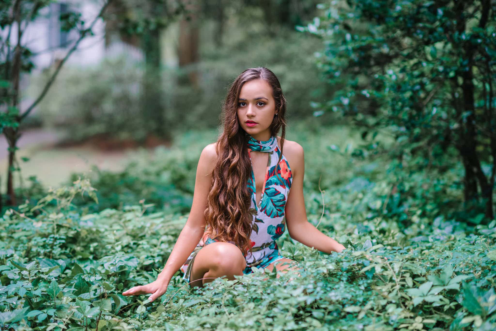 Vibrant outdoor portrait of a young woman with long wavy hair in a lush green garden setting, wearing a colorful floral dress, capturing natural beauty and effortless elegance in nature.