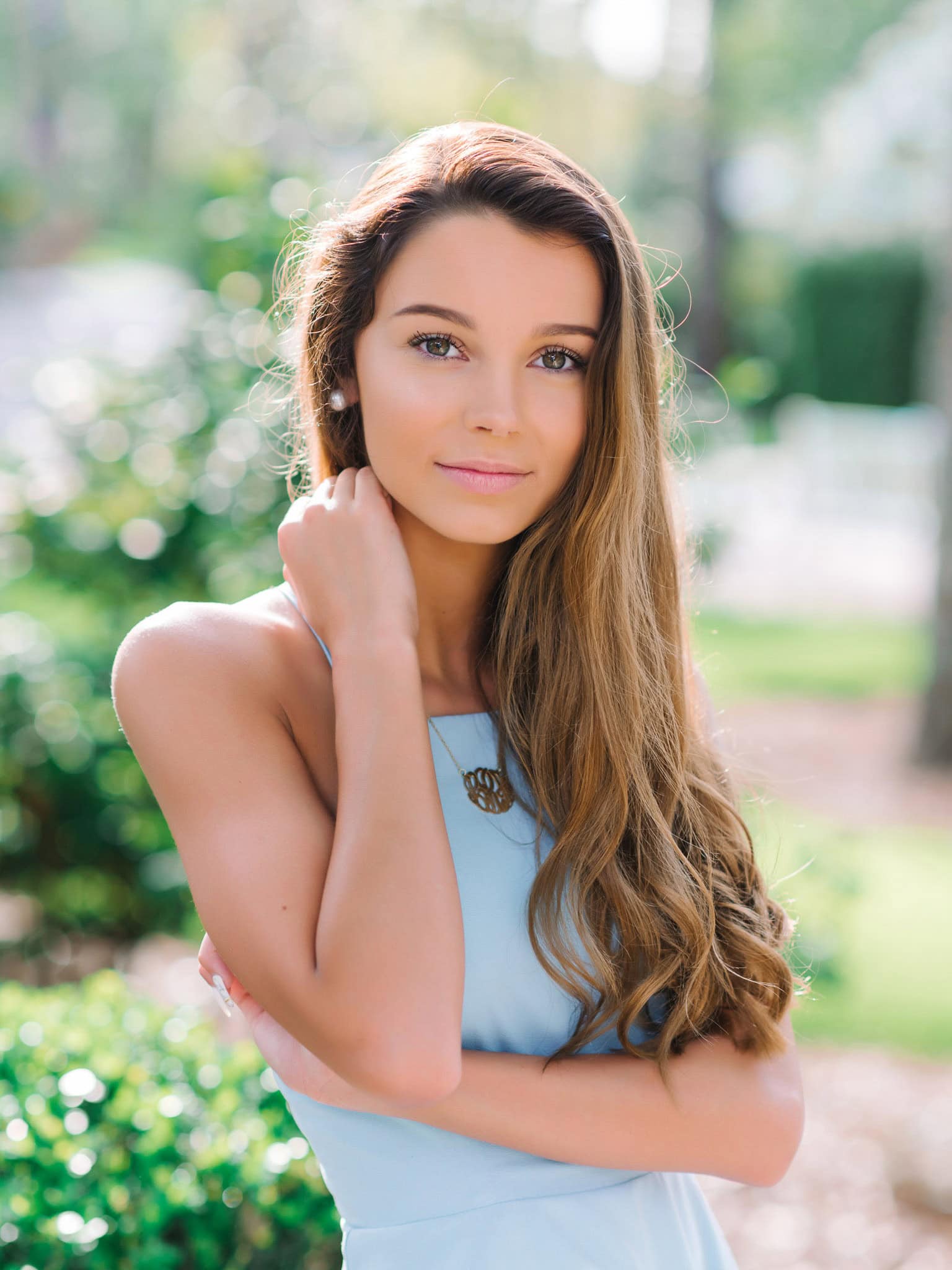 Beautiful young woman with long brown hair and blue eyes, outdoor portrait in natural sunlight, wearing a light blue dress, cheerful and elegant appearance, suitable for senior portraits or fashion photography.