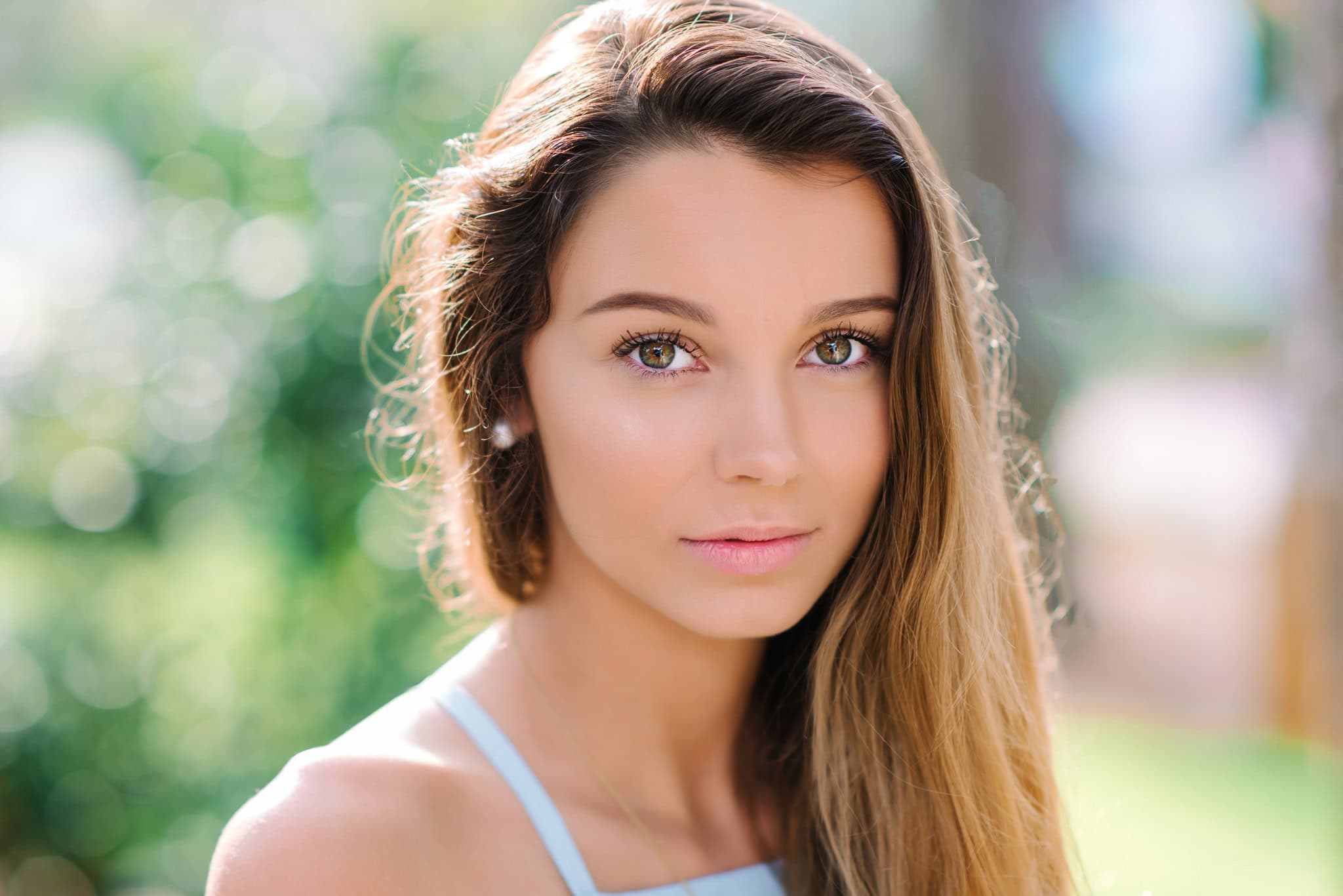 Stunning portrait of a young woman with long brown hair and bright hazel eyes, captured outdoors on a sunny day with a vibrant green blurred background.