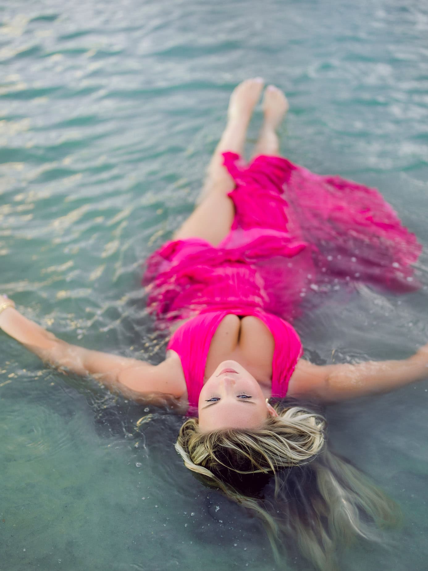 Vibrant woman in pink dress relaxing in clear ocean water during beach photoshoot, enjoying a serene moment in a family beach portrait setting.