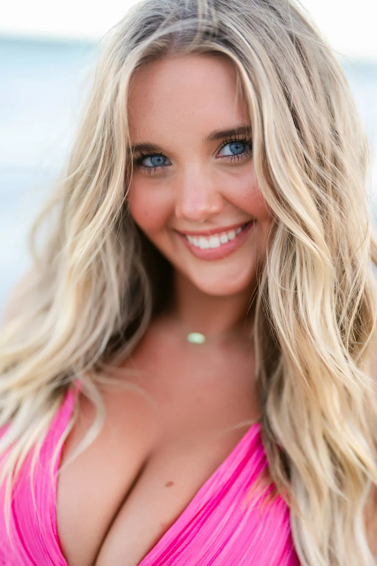 Bright blonde woman with blue eyes smiling in a pink dress, outdoor portrait, vibrant colors, professional photography for family beach portraits and senior photos.