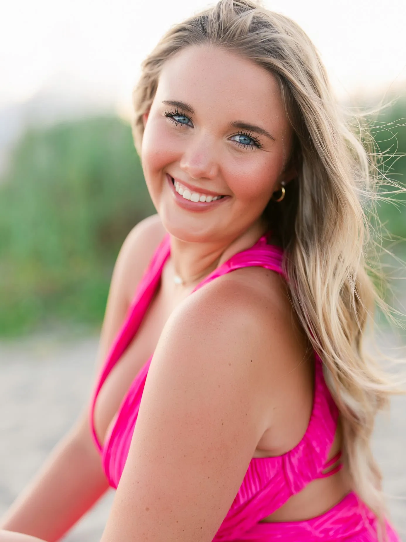 Bright smiling woman in a hot pink dress with blonde hair, outdoor portrait, natural sunlight, professional beach photography, joyful and vibrant lifestyle shot, close-up candid shot.
