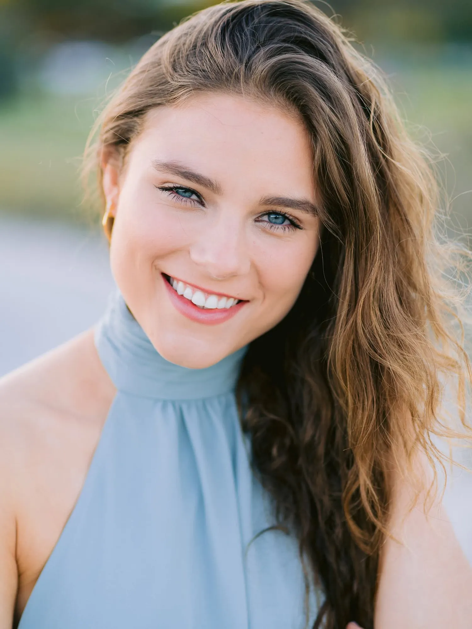 Bright smiling woman with blue eyes and wavy brown hair, wearing a light blue halter top, outdoors during golden hour, vibrant portrait.