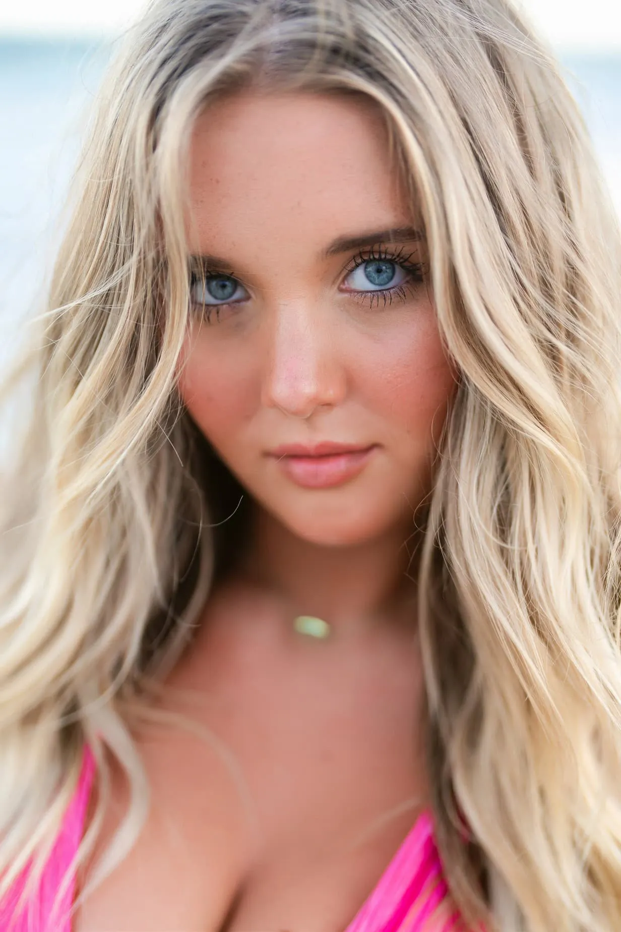 Close-up portrait of a young woman with striking blue eyes, blonde wavy hair, and natural makeup, wearing a pink swimsuit on the beach, captured by professional beach portrait photographer.
