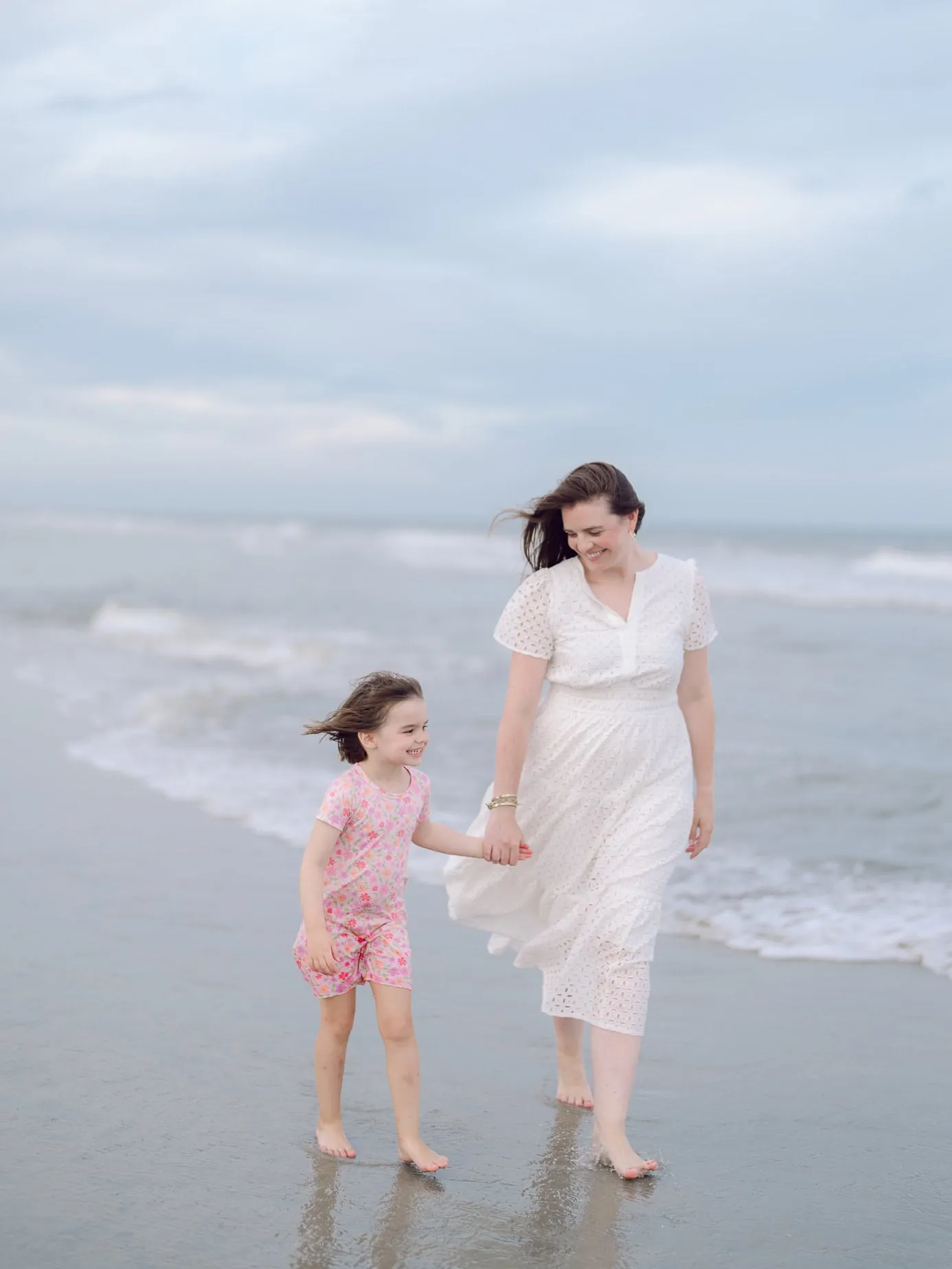 Mother and daughter walking hand in hand along the shoreline during their family beach session