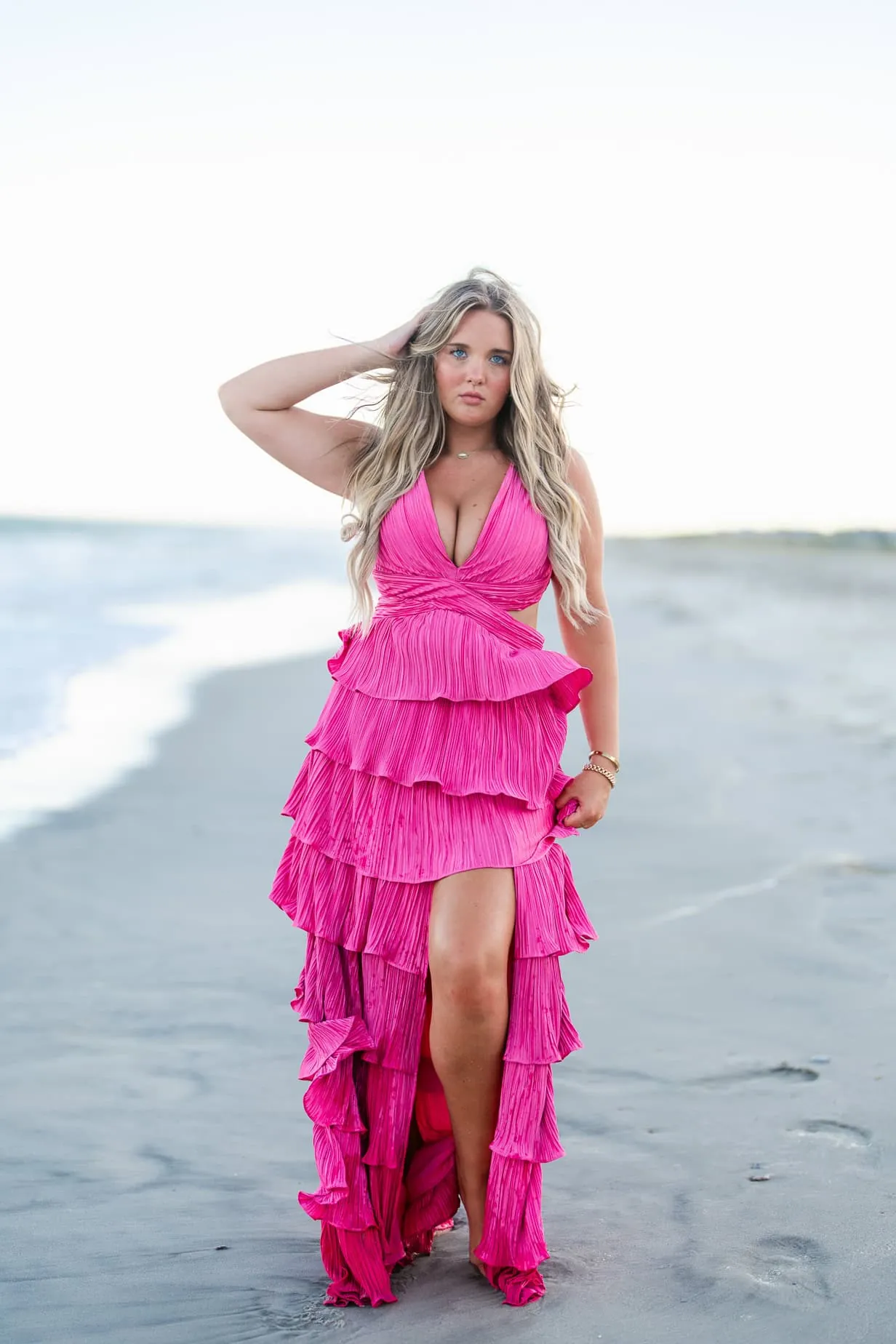 Stunning woman in a vibrant pink ruffled dress standing on the beach during sunset, emphasizing high-end fashion photography for beachwear and personal style shots.