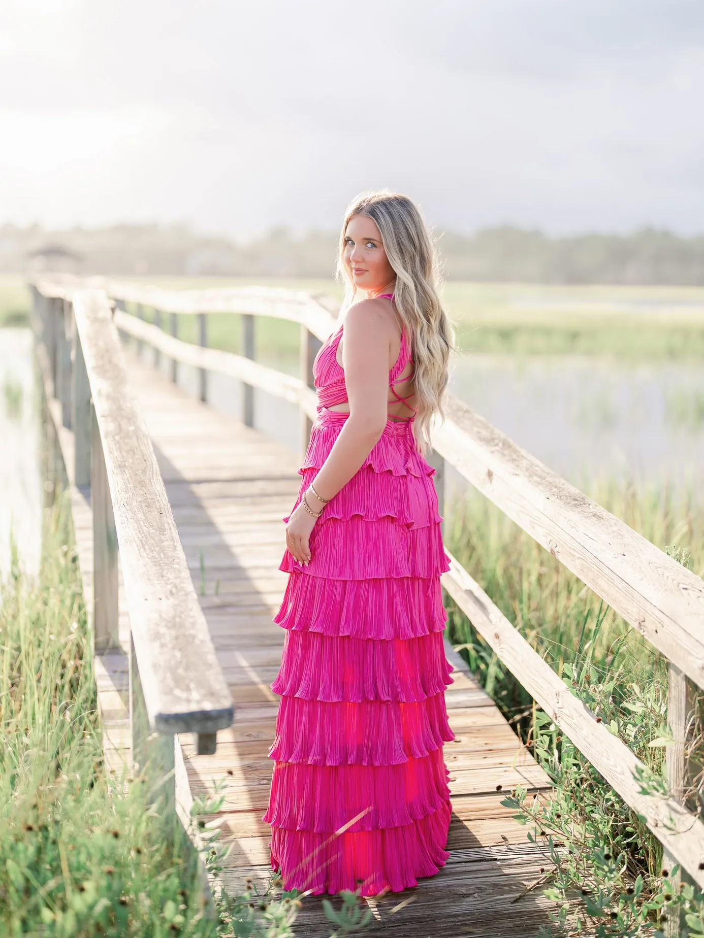 Stunning young woman in a bright pink ruffled maxi dress standing on a wooden bridge in a natural outdoor setting with water and greenery, perfect for studio senior portraits.