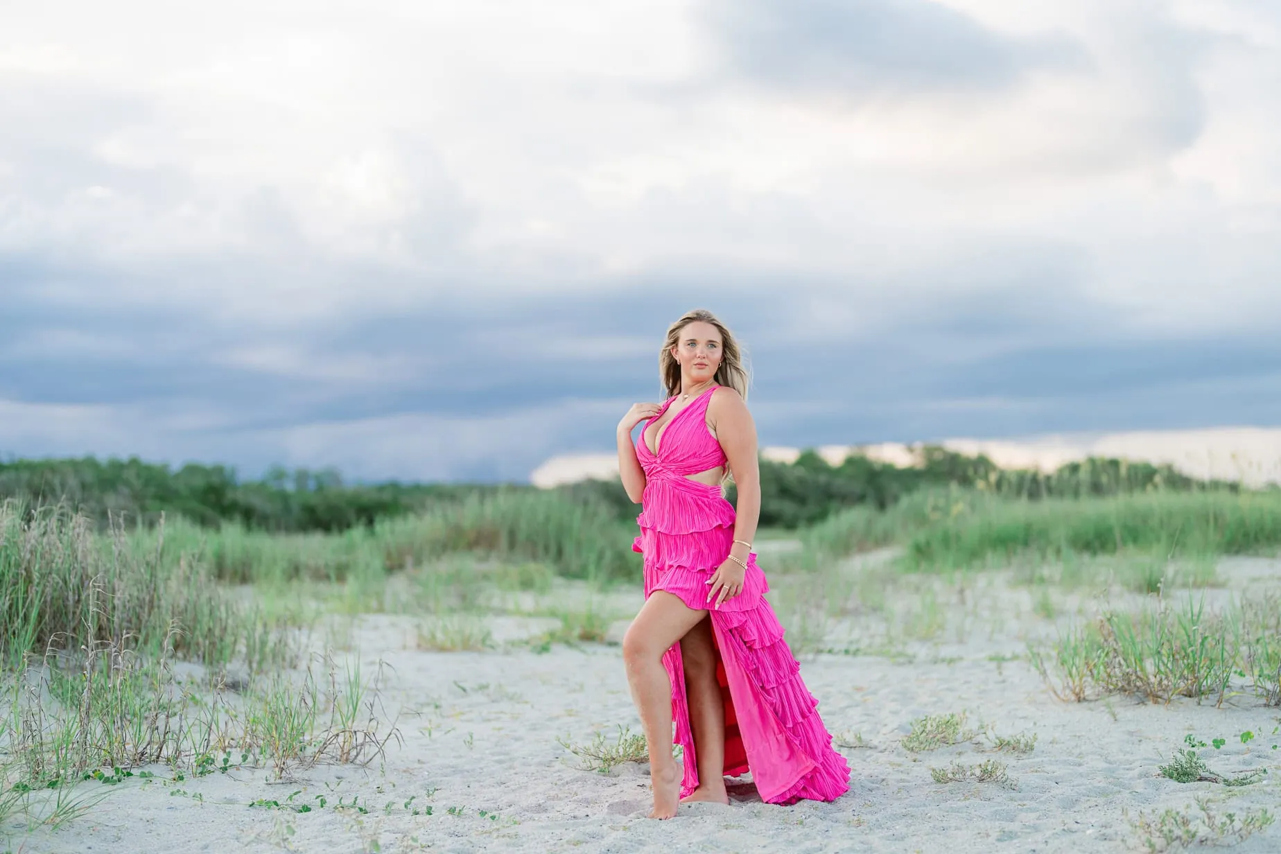 Vibrant beach portrait of a high school senior in a pink dress with lush green dunes and a cloudy sky, perfect for family beach photography and senior portraits on the sand.