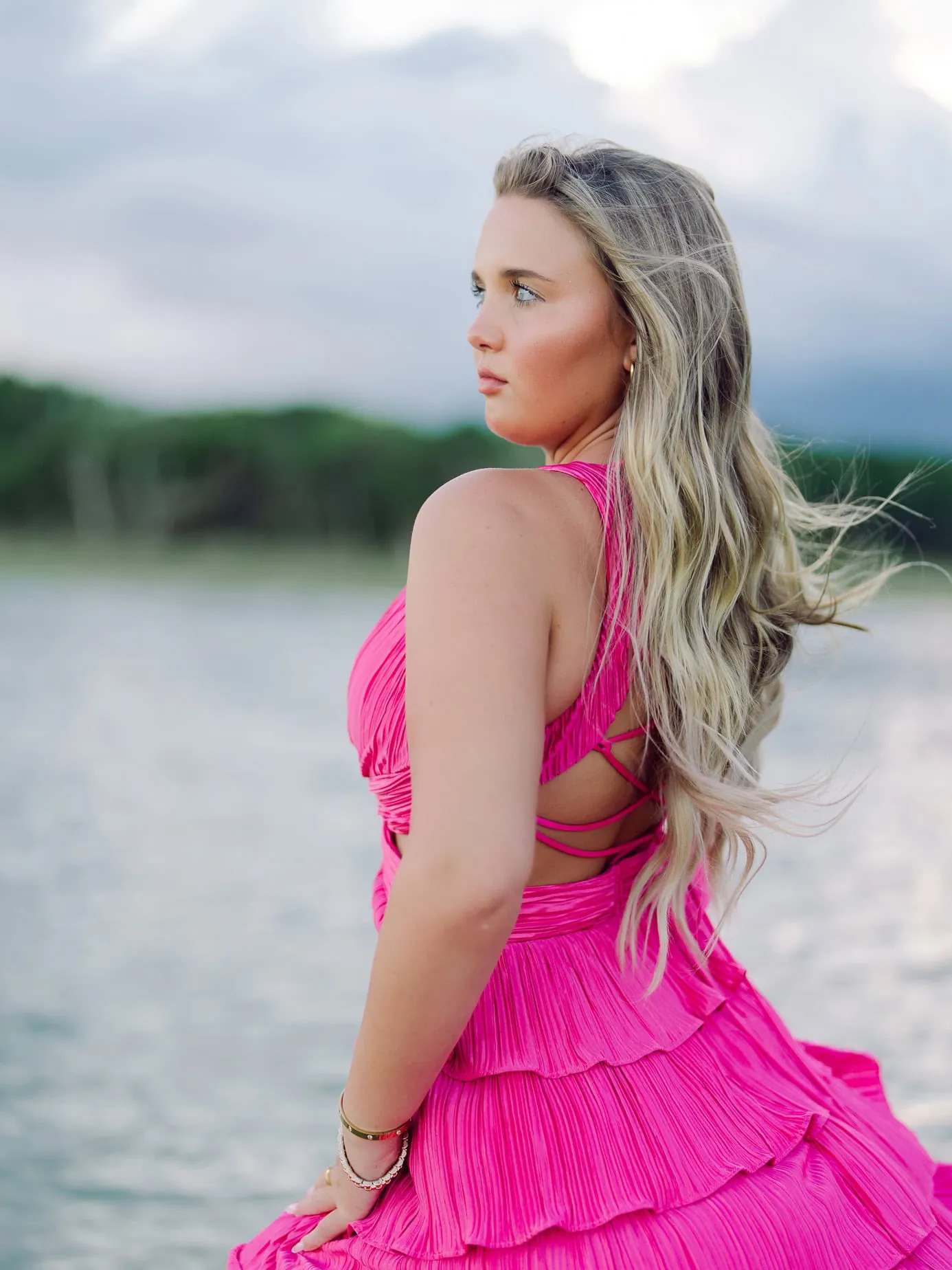 Vibrant fashion portrait of a young woman in a bright pink dress with flowing blonde hair, standing near a serene water body with lush greenery in the background, during outdoor photography session.