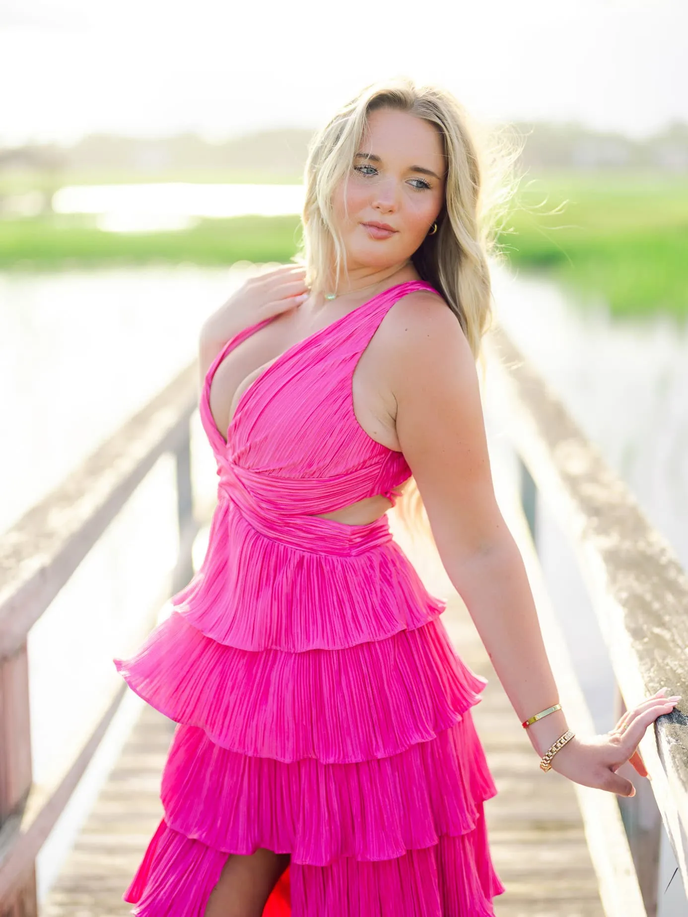 Vibrant pink tiered dress on young woman posing on a wooden bridge over water, outdoor portrait with natural backlit glow, colorful fashion and radiant expression, perfect for fashion or lifestyle photography.