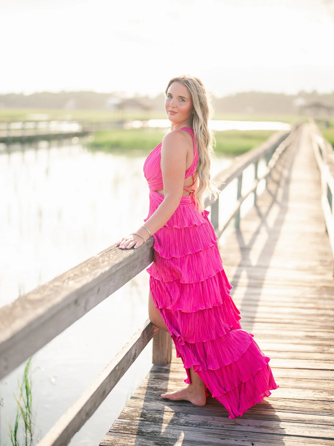 Vibrant pink tiered dress worn by a woman on a wooden dock overlooking a serene marsh during golden hour.