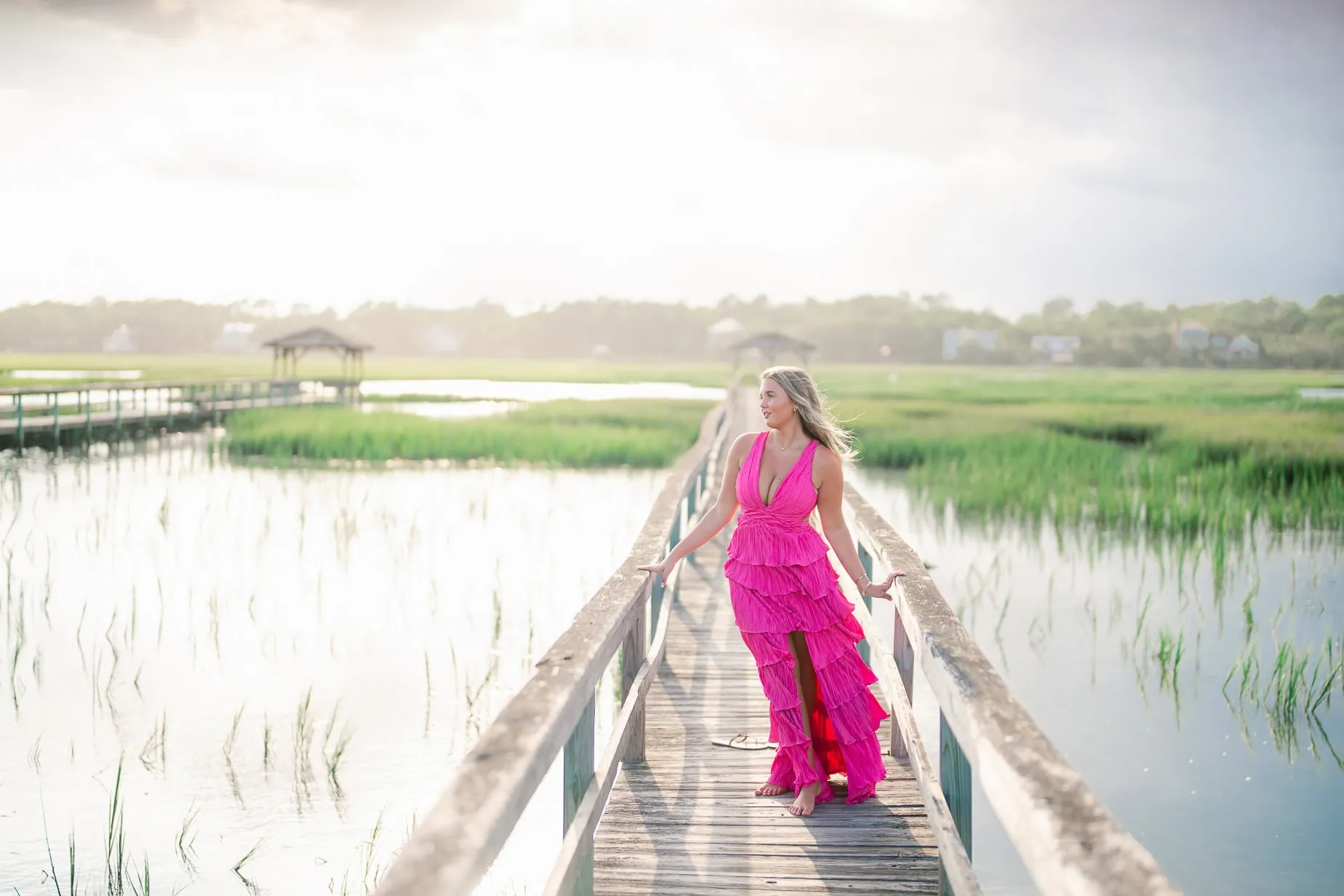 Vibrant woman in a bright pink层 dress walking along a wooden dock over a marshy landscape, featuring lush green marsh grasses and calm water, capturing a serene portrait outdoors.