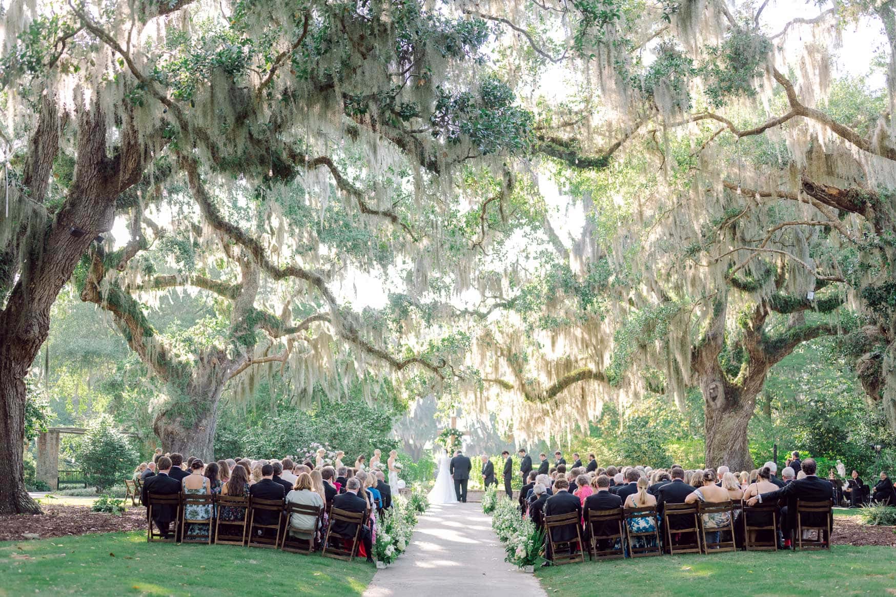 Lush outdoor wedding ceremony under large moss-draped trees with guests seated and couple exchanging vows in natural light setting.