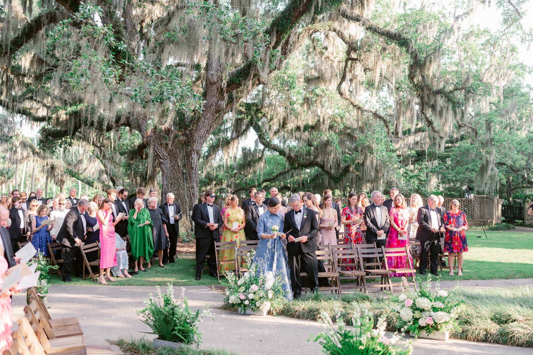 Vibrant outdoor wedding ceremony under a sprawling moss-draped tree with guests in formal attire, capturing family and friends celebrating love in a lush garden setting.