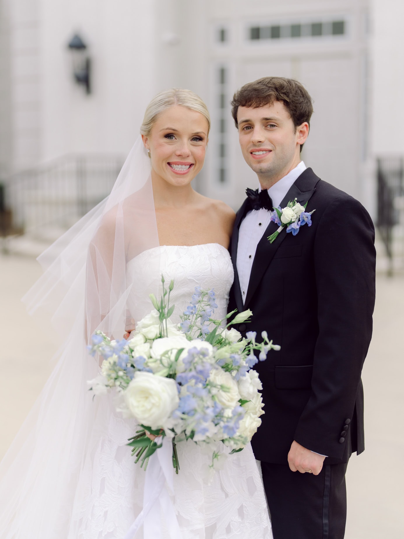 Elegant wedding couple standing outdoors in front of a scenic coastal house, dressed in traditional wedding attire with floral accessories, capturing a joyful moment of their special day.