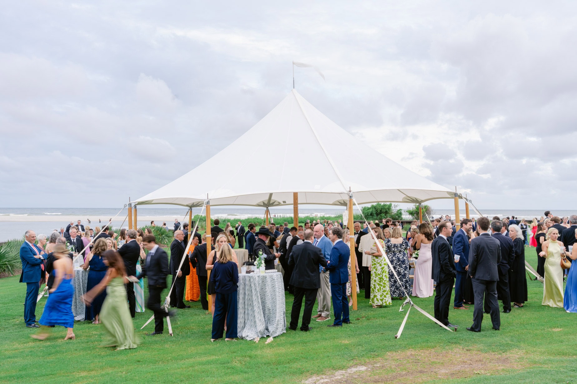 A lively outdoor wedding reception under a large white tent on a lush green lawn next to the beach with guests mingling and enjoying the ocean view.