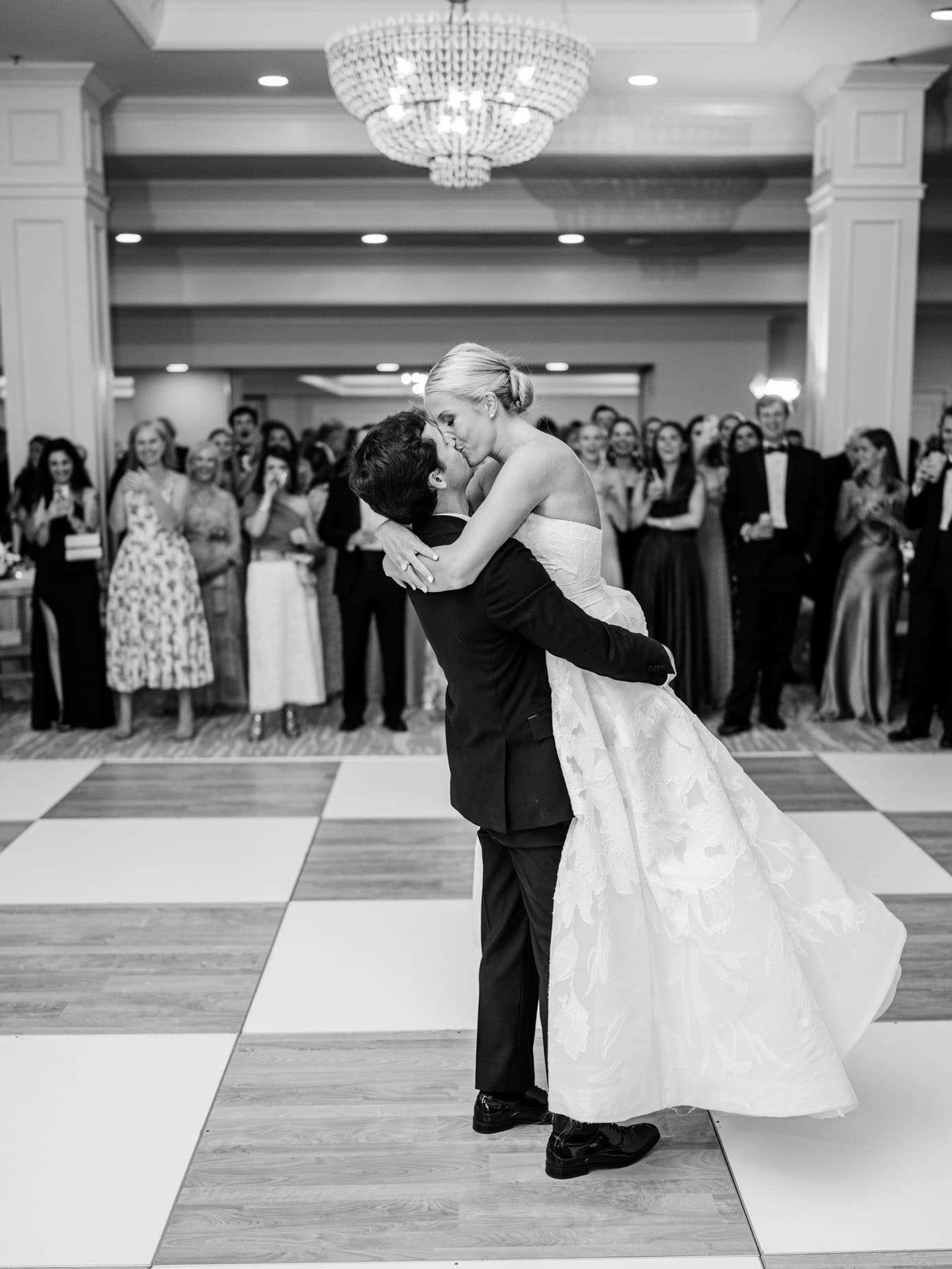Dancing couple during wedding reception with guests watching, black and white wedding photo, romantic first dance, elegant wedding celebration, timeless wedding photography.