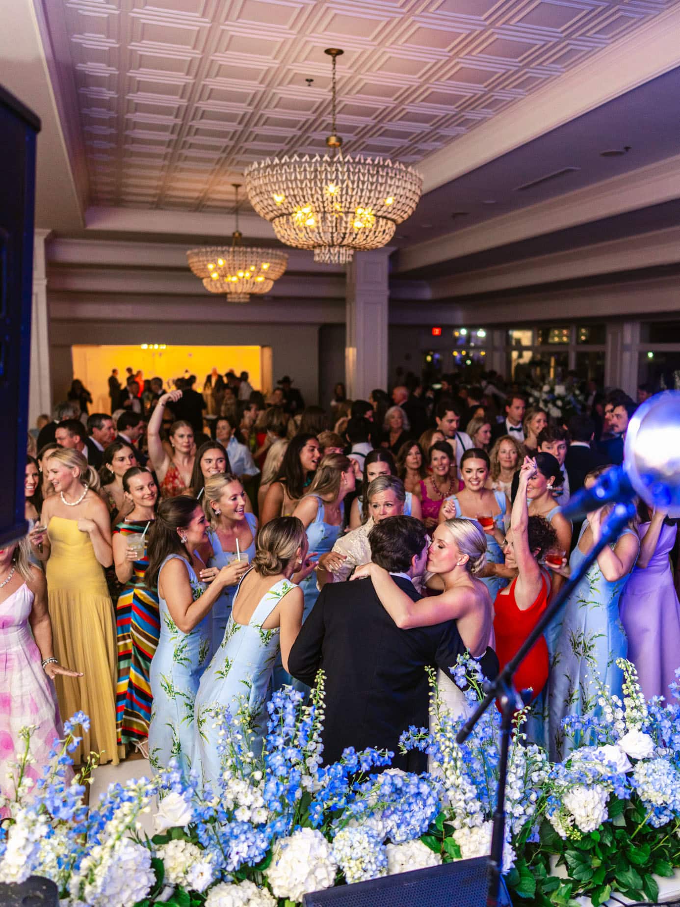 Dancing wedding guests at reception with colorful attire, joyful celebration, and floral decorations; capturing lively moments of wedding party and family at an elegant indoor event.