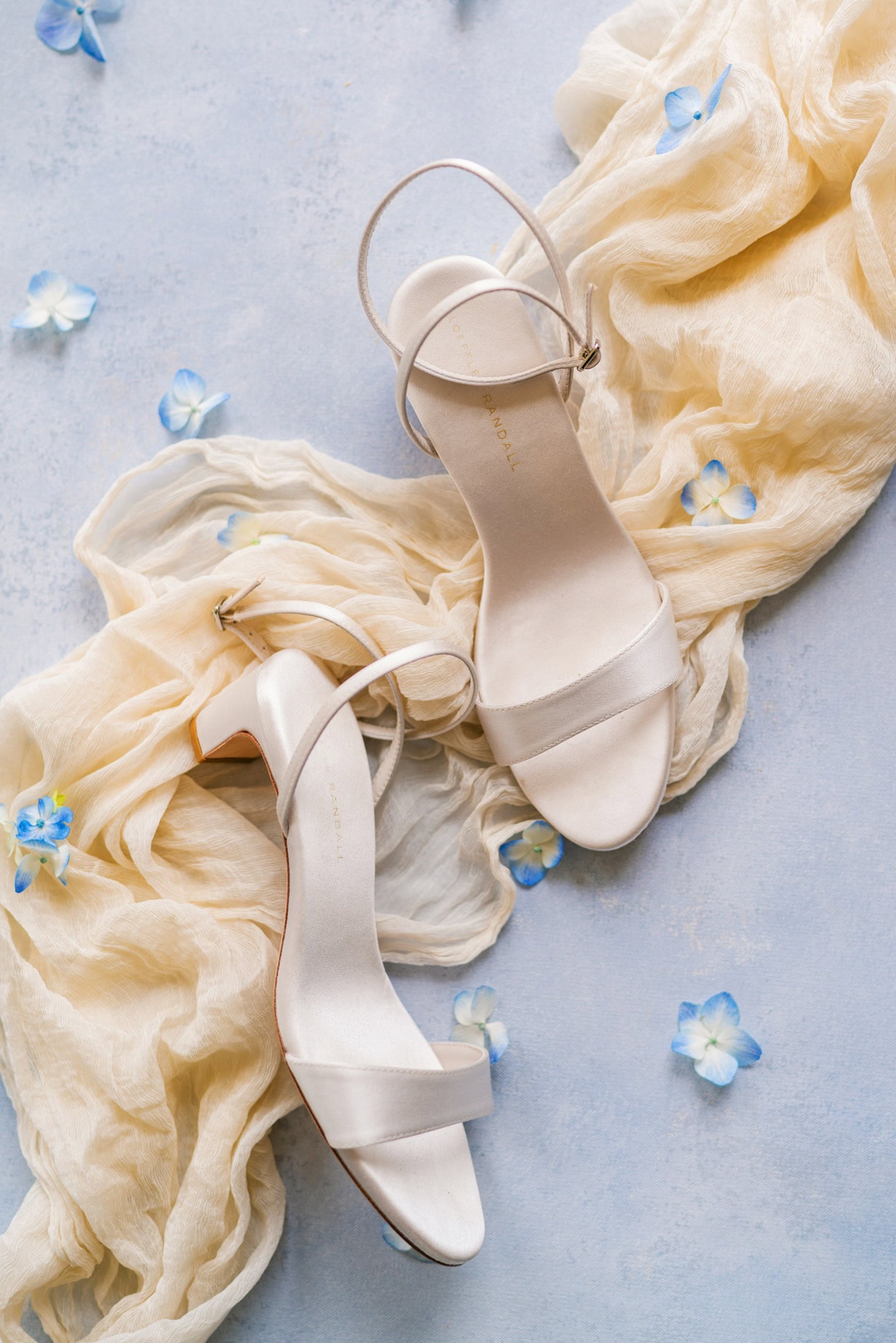 Elegant white wedding heels on soft yellow fabric with blue flower accents, perfect for wedding photography and bridal portrait sessions.
