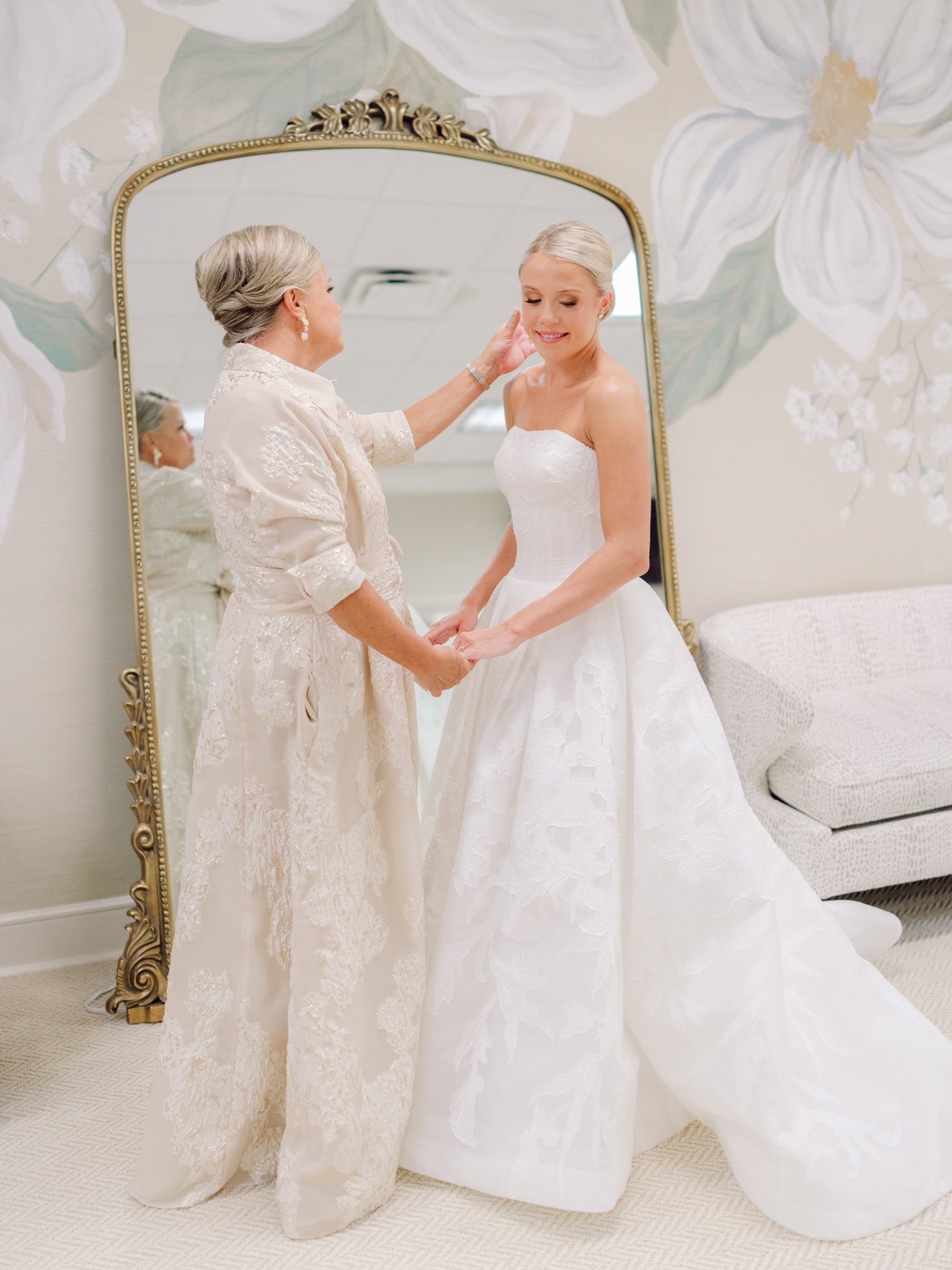 Elegant wedding bride in a strapless lace gown with her mother getting ready, touching her face in a stylish bridal prep moment, with a large mirror and floral wall art in the background.