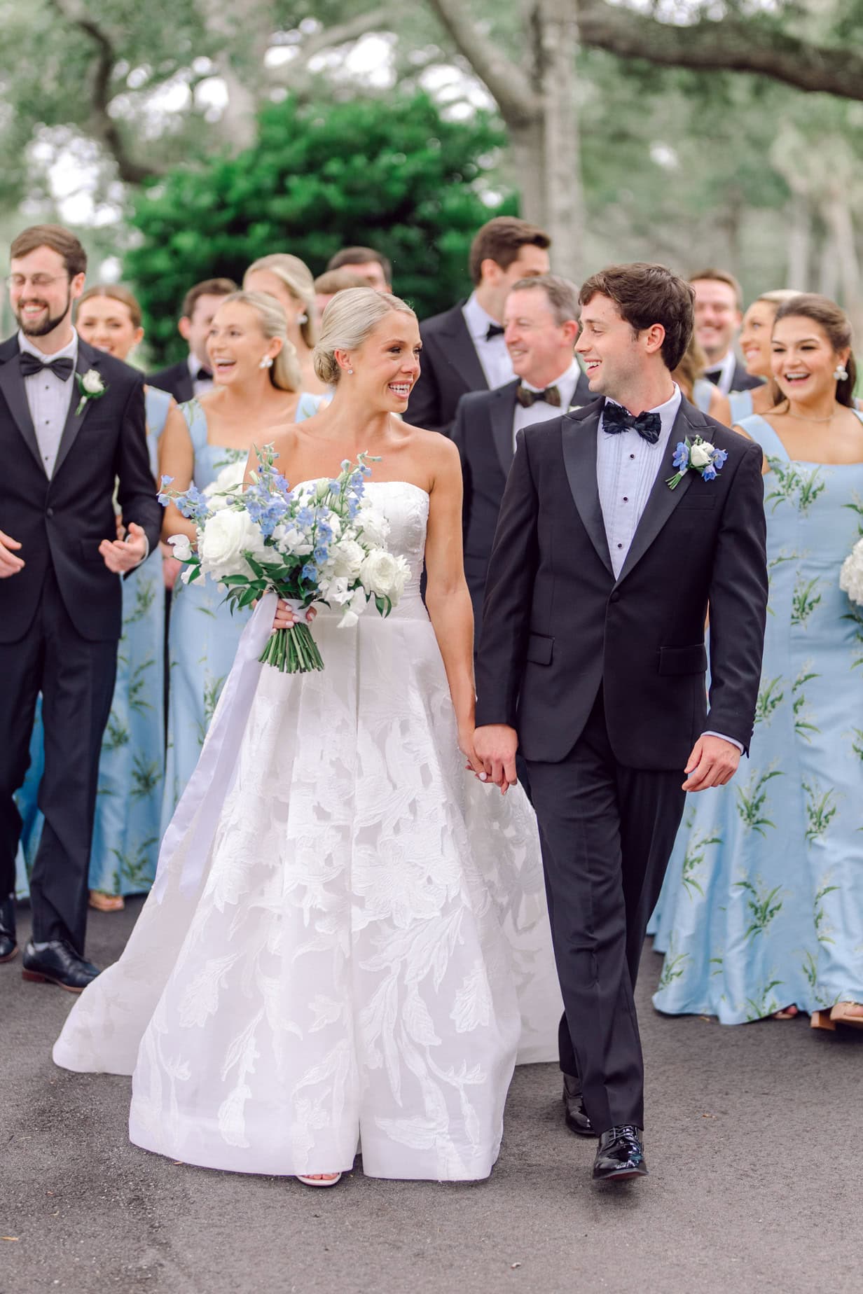 Elegant bride and groom holding hands during wedding celebration with bridal party in background, outdoor wedding photography capturing happiness and love among family and friends.