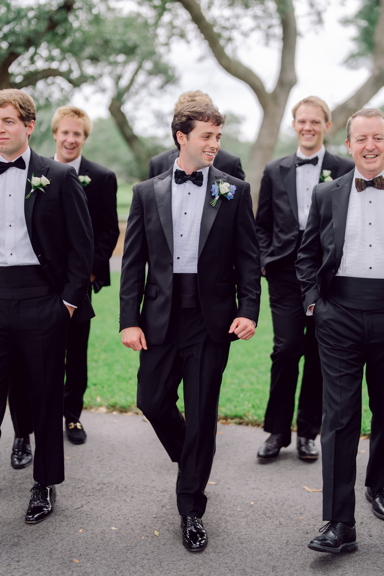 Elegant groom walking with groomsmen in tuxedos at outdoor wedding, stylish wedding party, formal wedding attire, joyful wedding celebration, classic black-tie wedding photography.