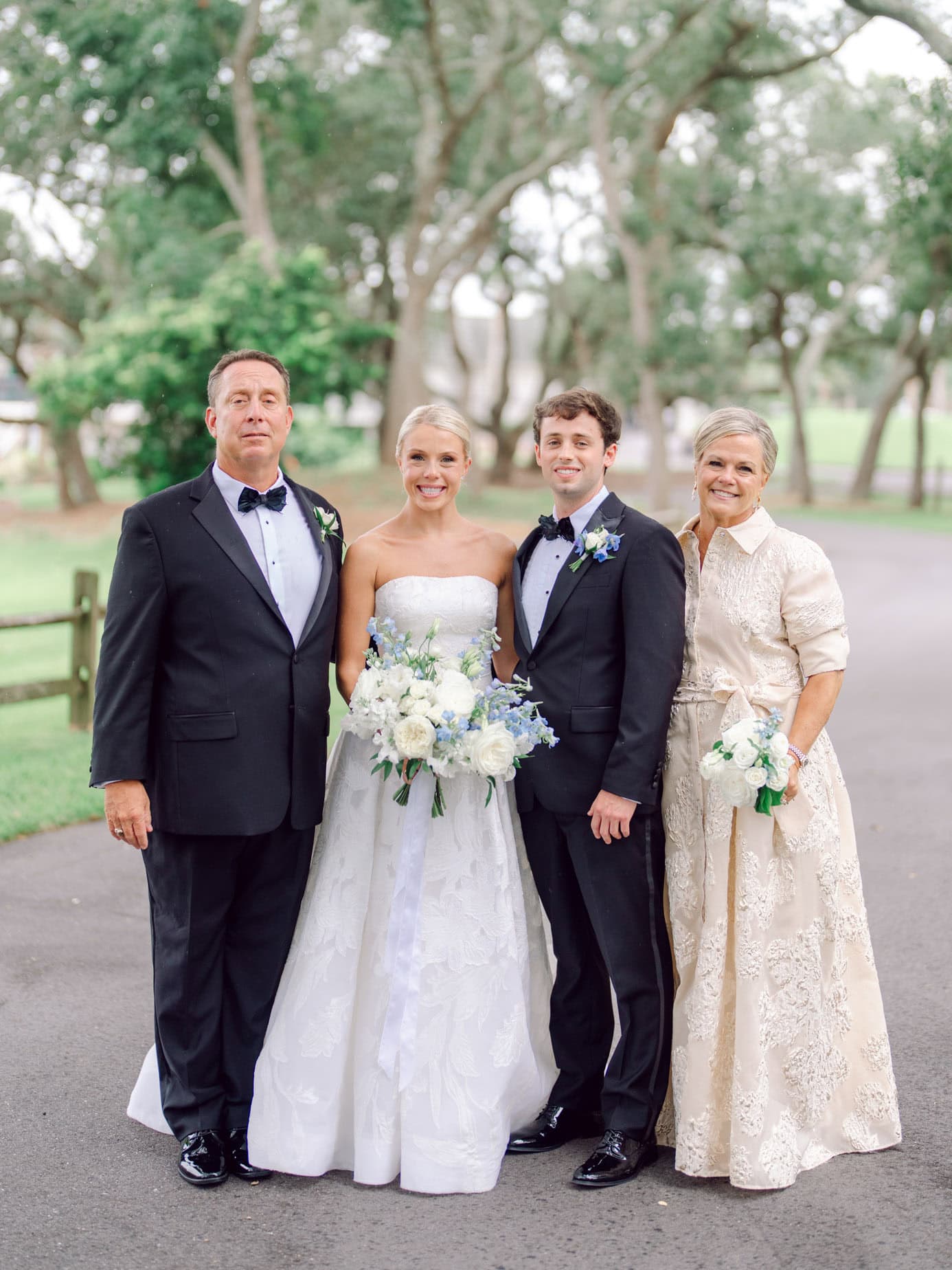 Elegant wedding portrait of a bride, groom, and their parents outdoors, featuring beautiful garden scenery, formal attire, and joyful expressions capturing a special moment.