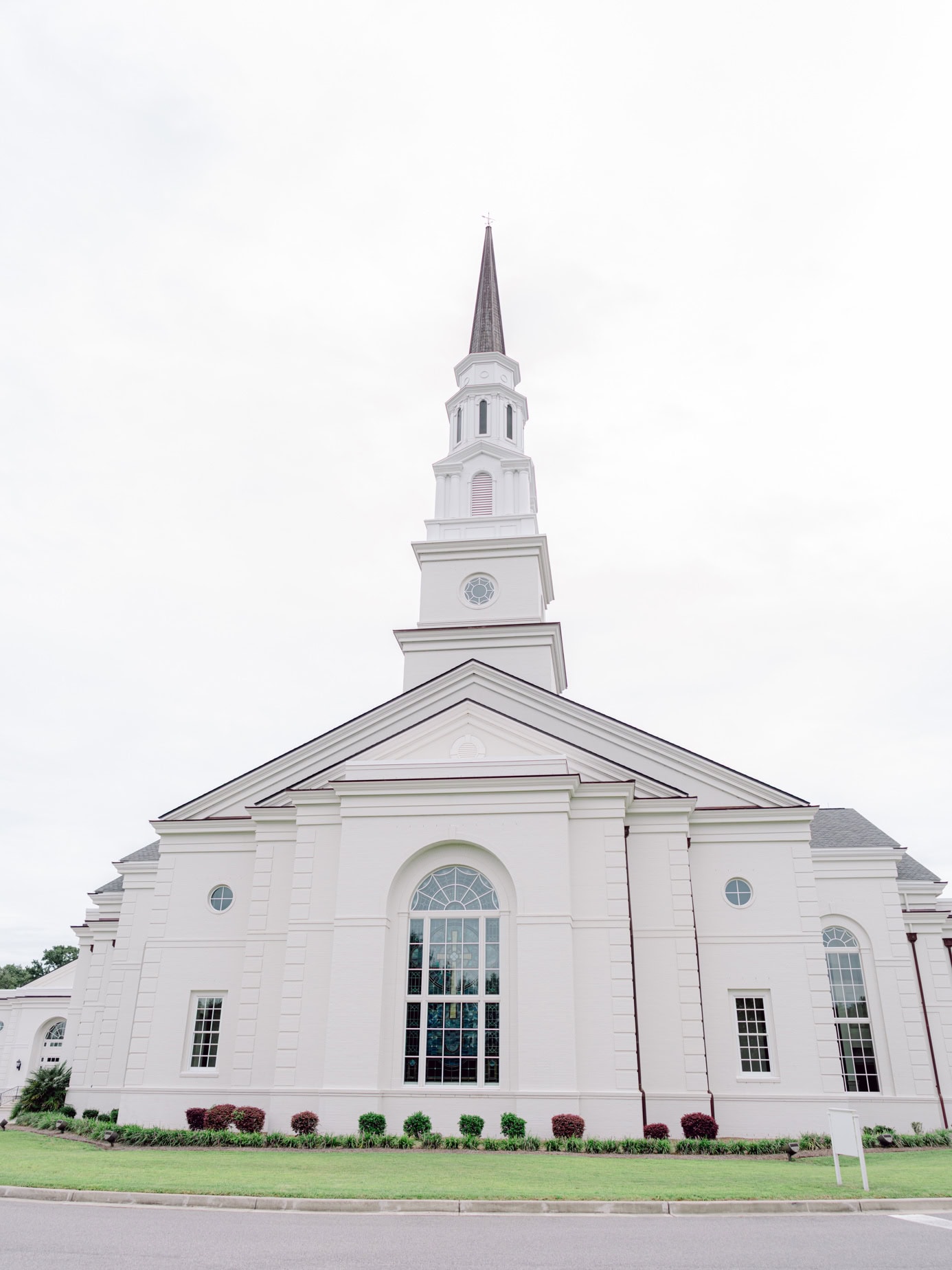 Stunning white church with tall steeple and large stained glass windows, ideal location for wedding photography and special events. Beautifully maintained exterior with manicured lawn and vibrant landscaping.