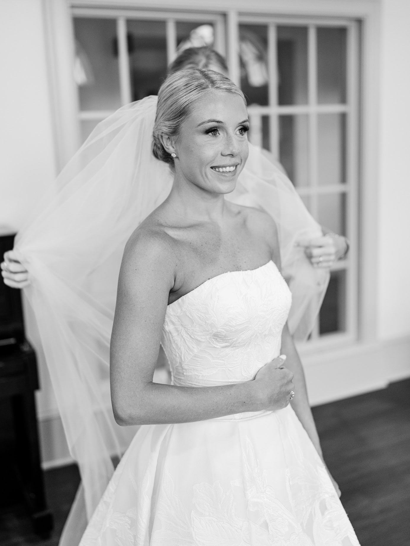 Elegant bride preparing for her wedding day, smiling confidently in strapless wedding gown with veil, captured in a candid black and white photo.