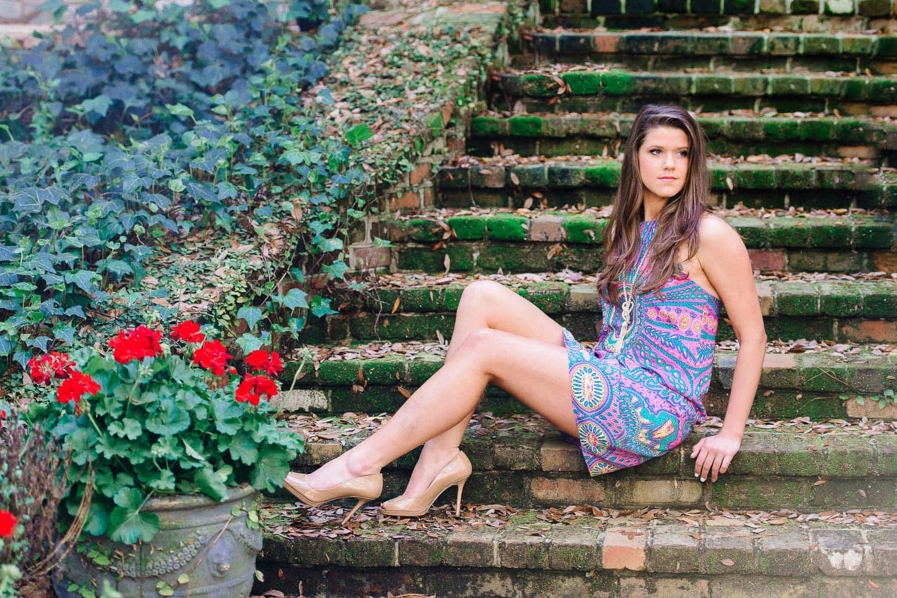 Vibrant woman in colorful summer dress sitting on mossy brick stairs with red flowers and lush greenery, outdoor portrait, natural light, lifestyle photography, senior photos, garden or park setting.