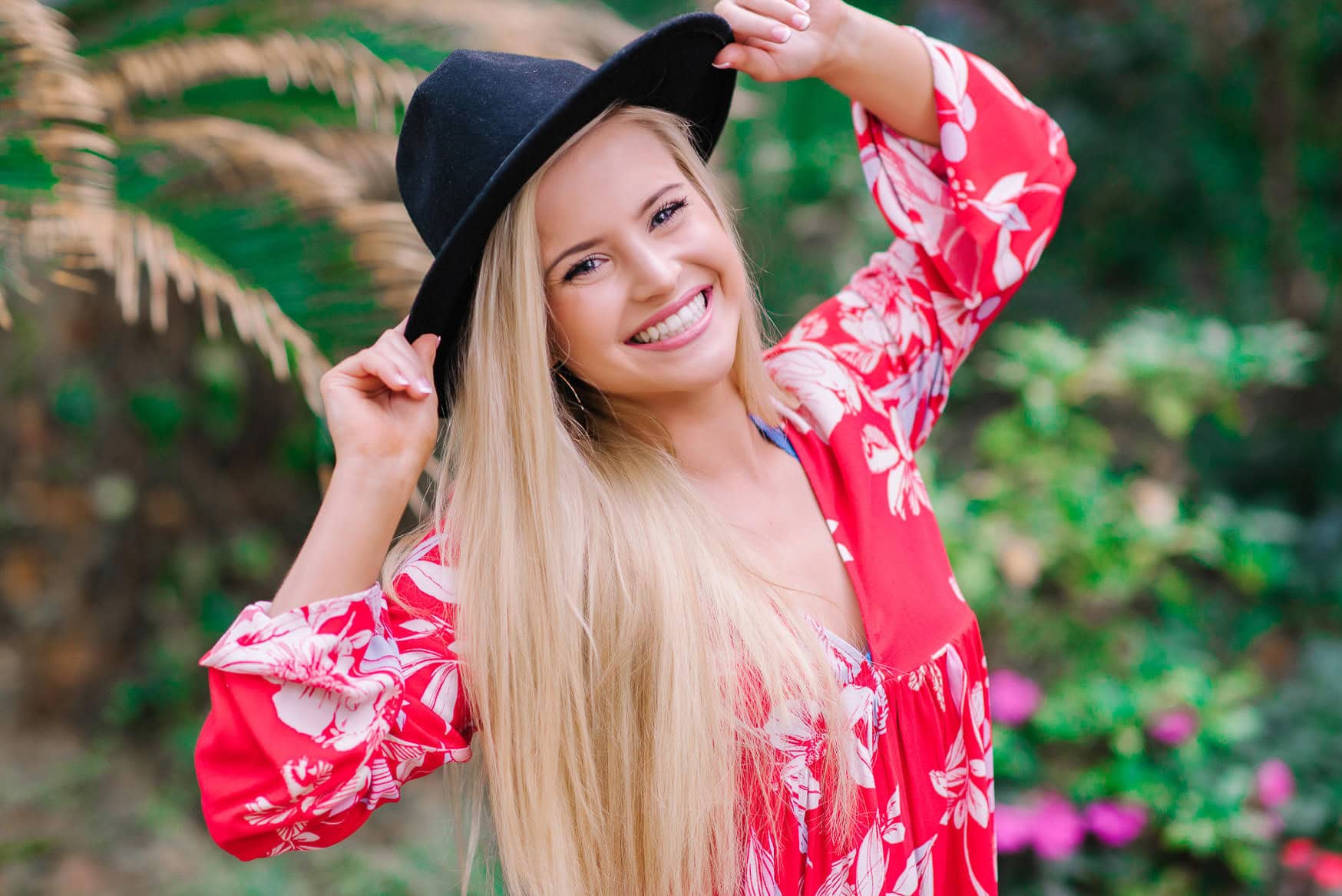 Youthful blonde woman in a vibrant red floral dress smiling outdoors while adjusting her black hat, surrounded by lush greenery and blooming pink flowers.