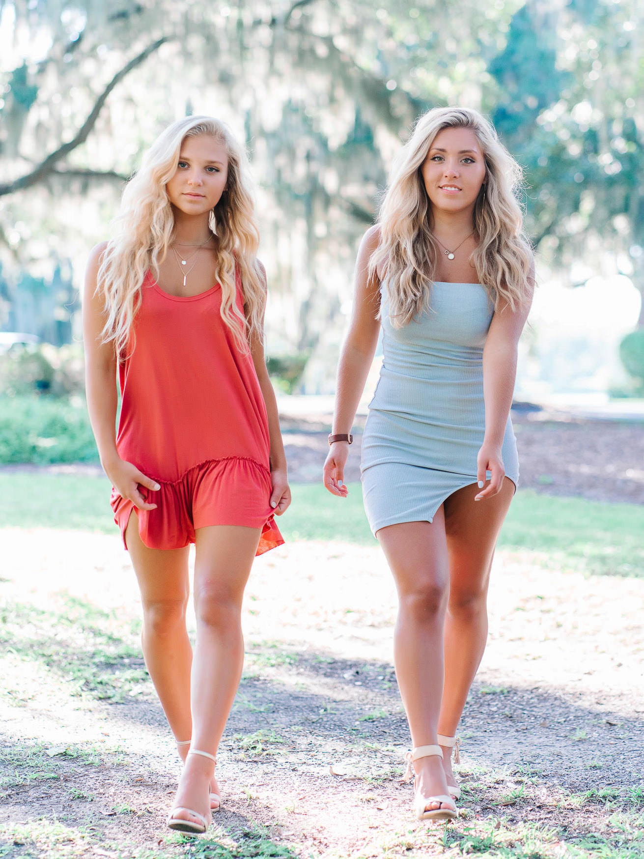 Vibrant outdoor portrait of two young women with long wavy blonde hair walking in a sunlit park, showcasing family beach photos and senior portraits with natural lighting and scenic background.