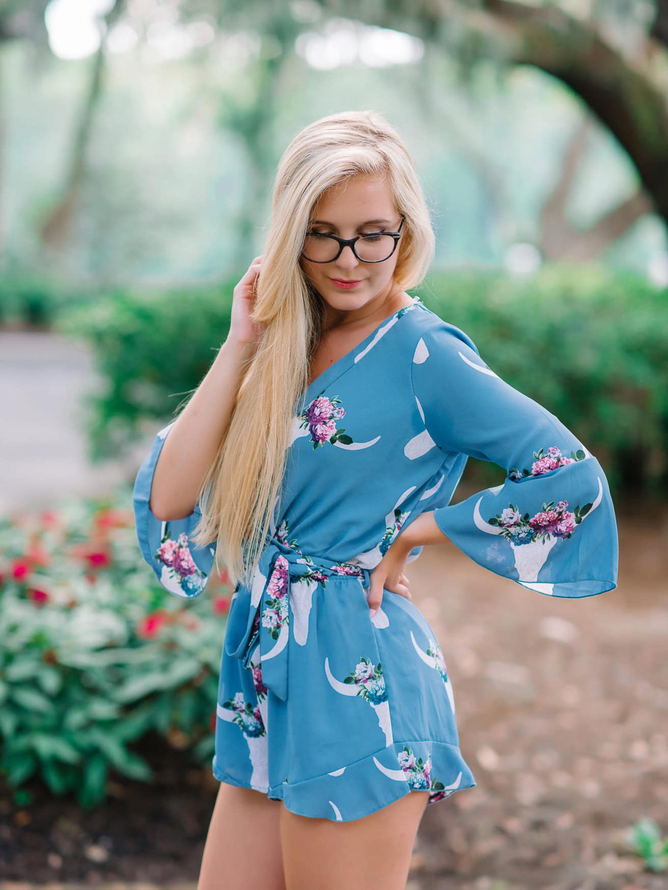 Elegant young woman in floral blue romper with long blonde hair and glasses, outdoor garden setting, casual fashion portrait.