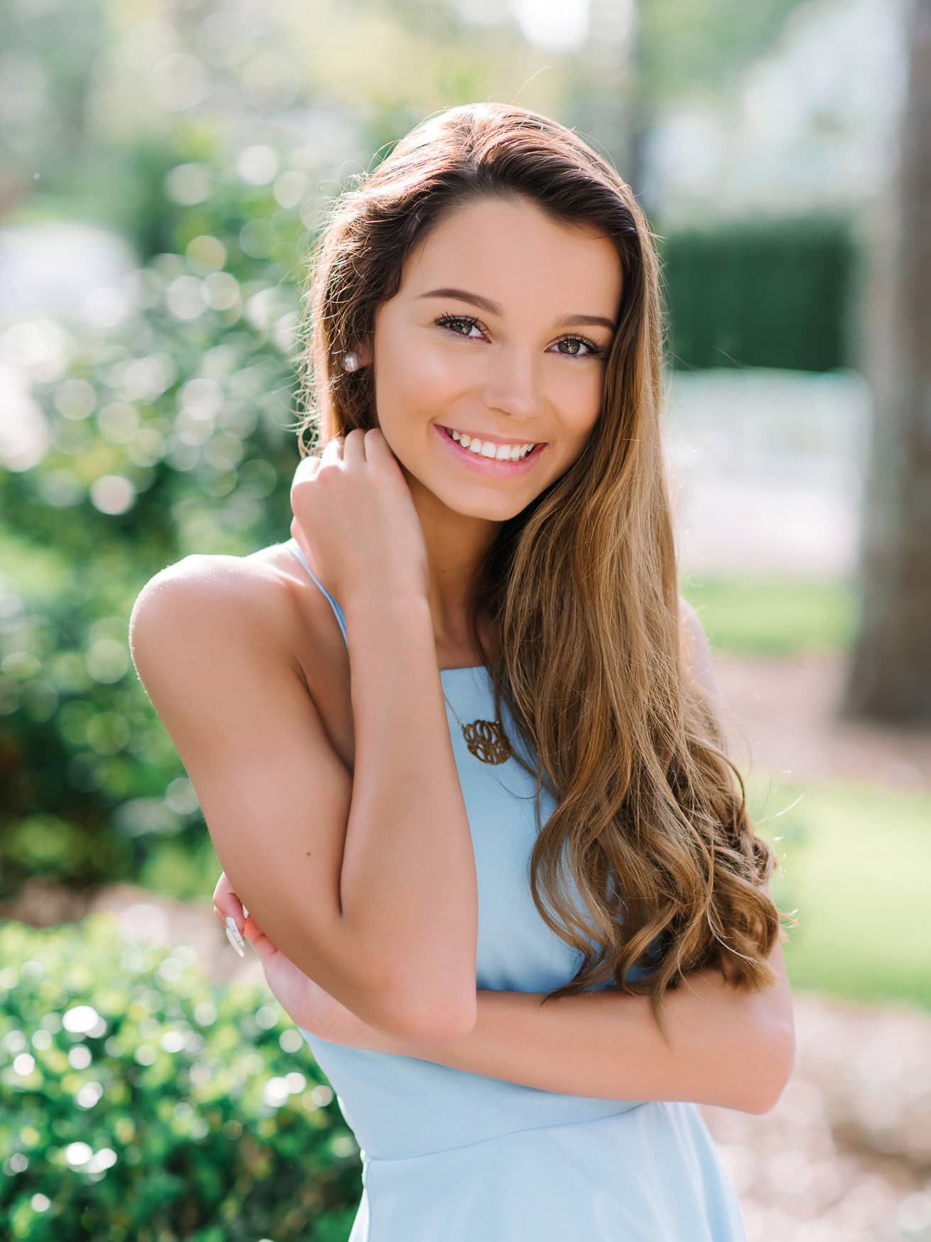 Bright, happy teenage girl with long, wavy brown hair, wearing a light blue dress and a gold necklace, smiling outdoors during a sunny day.