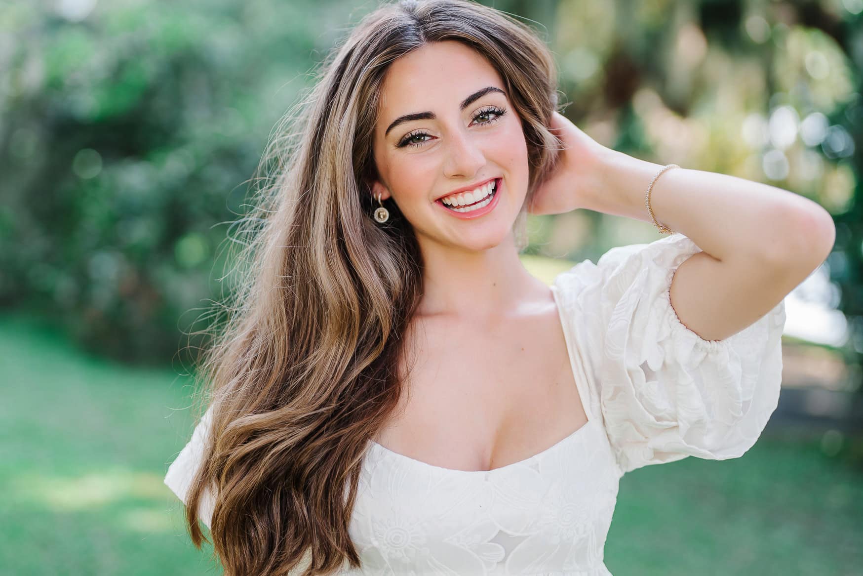 Bright young woman with long wavy hair, smiling confidently outdoors during daytime; perfect for senior portraits, wedding photography, or family beach sessions.