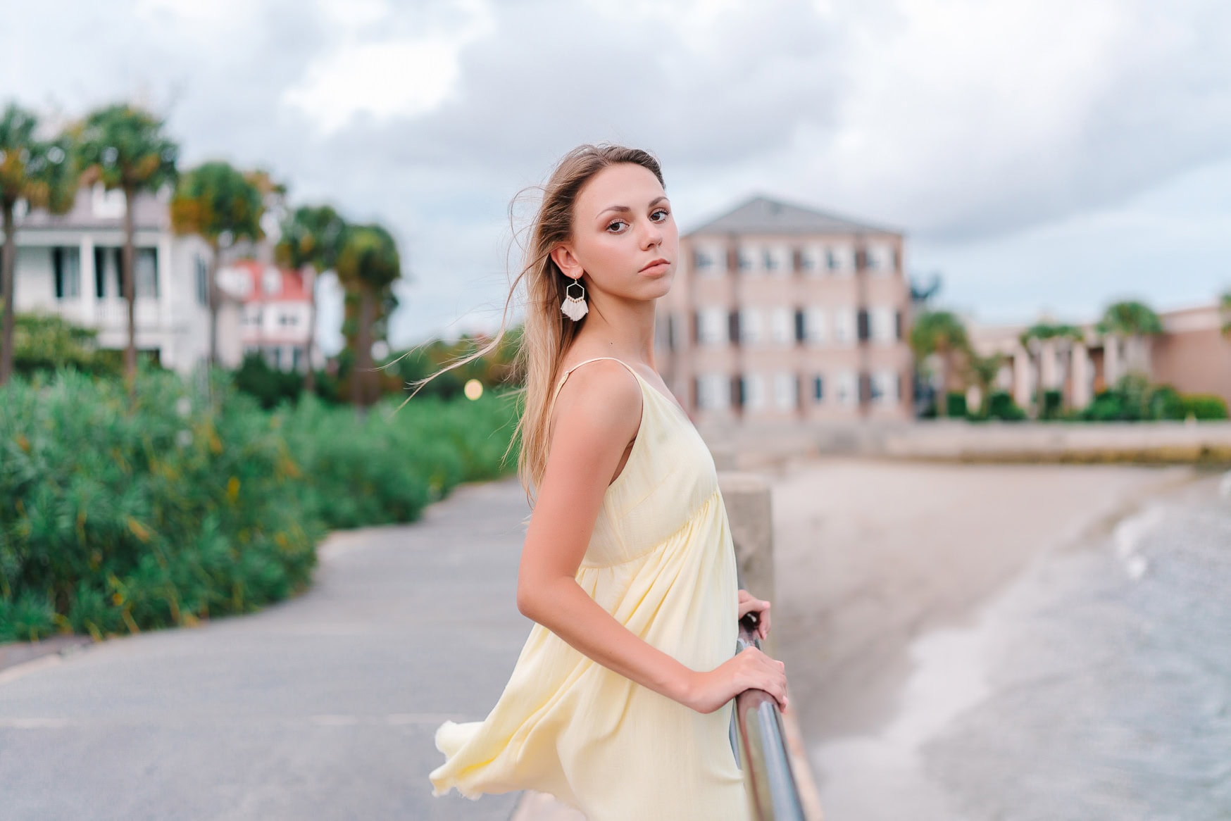 Stunning young woman in yellow dress posing on a beach full of greenery and colorful houses, capturing a serene coastal photography scene.