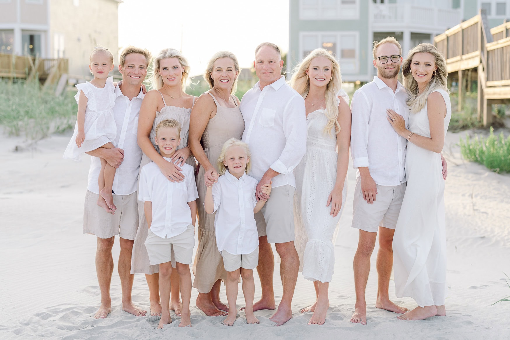 Family beach portrait featuring multiple generations, all dressed in white, standing on sandy shore with beach houses in the background. Perfect for family photography sessions and seaside wedding photos.