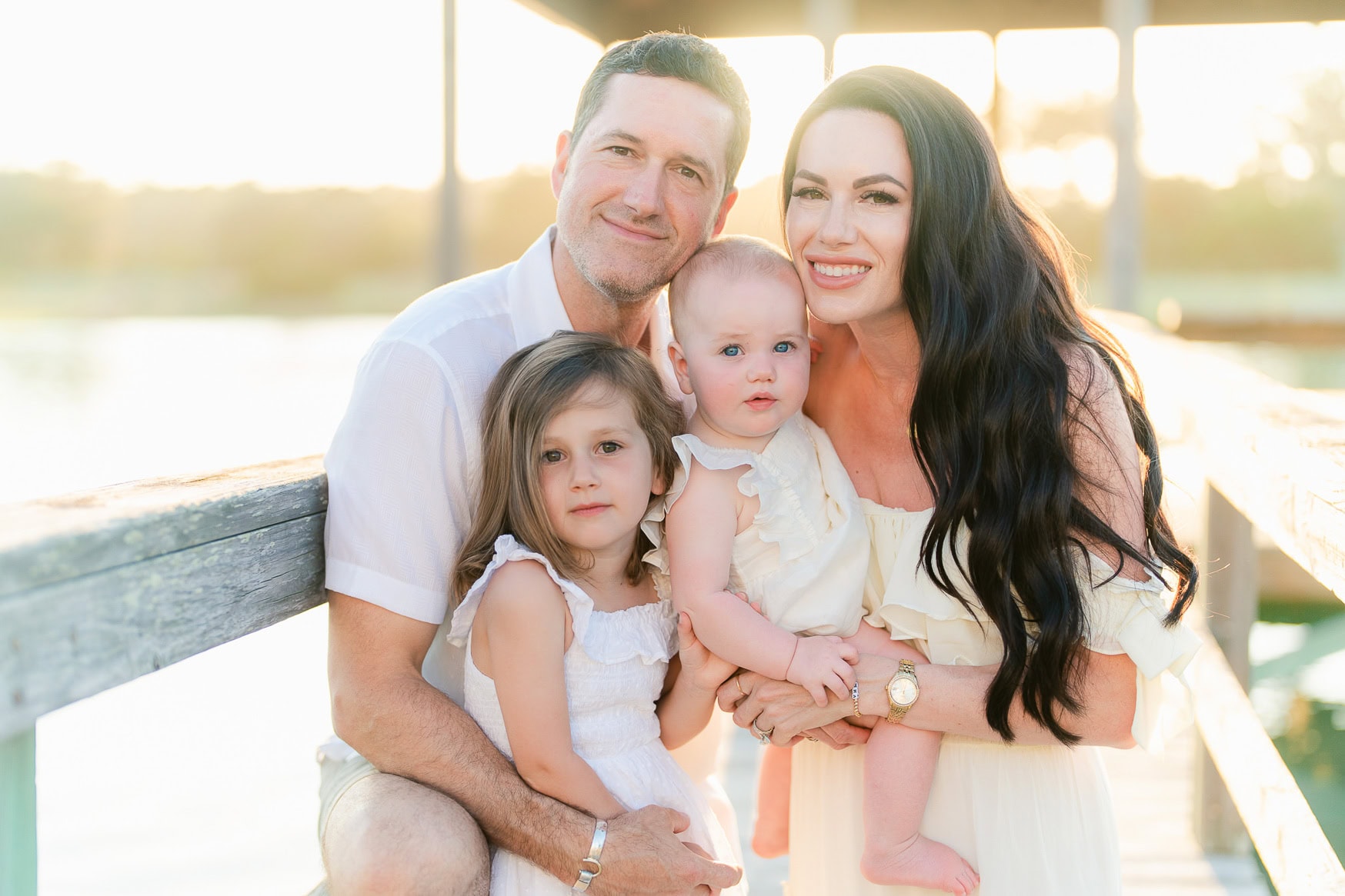Happy family portrait at the beach, parents with two young daughters, sunlit sunset background, capturing joyful outdoor family photography.