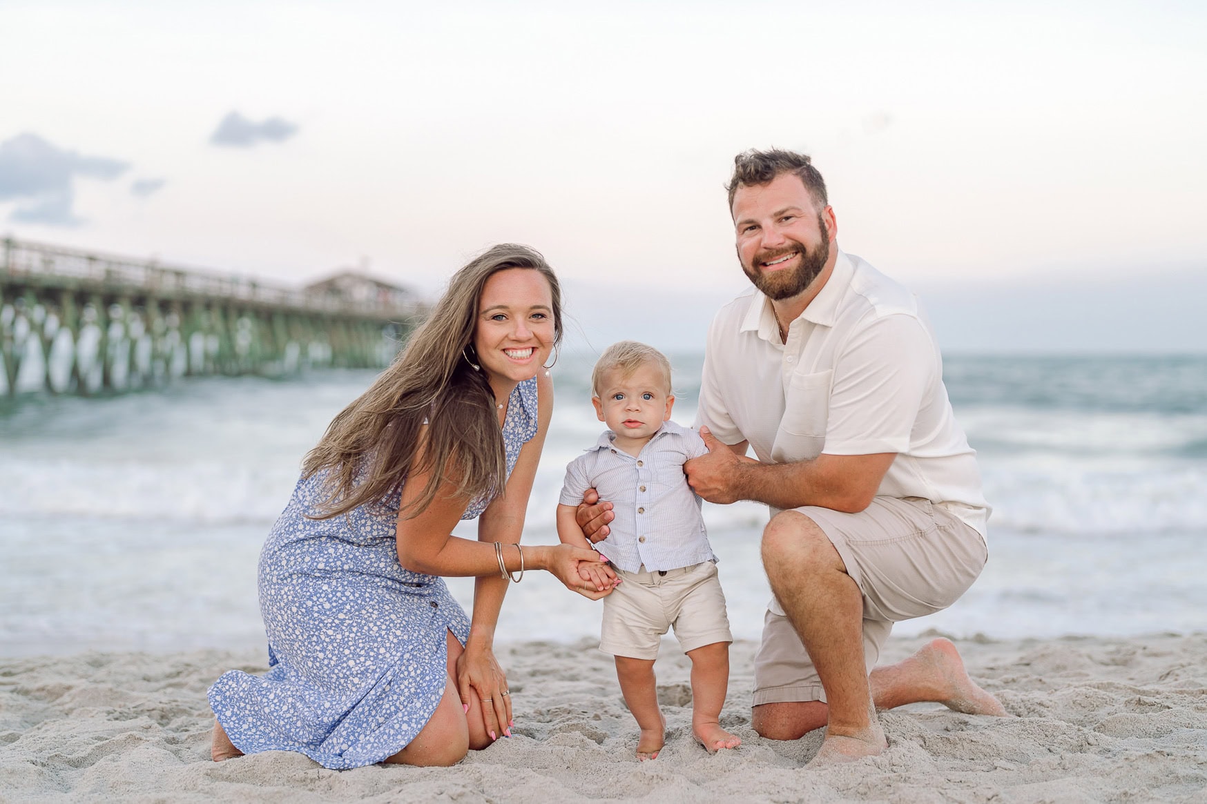 Playful family portrait on the beach with parents and young son, capturing joyful moments during sunset with ocean and pier in the background, perfect for beach family photography.