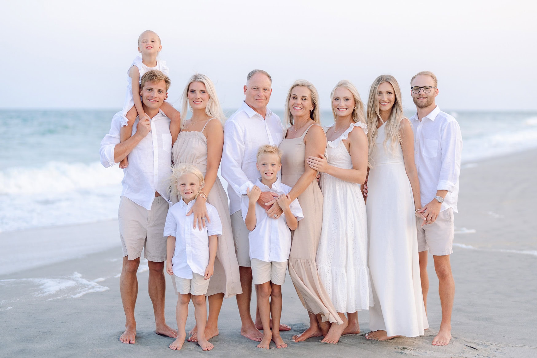Family beach portrait during sunset with everyone dressed in white, smiling happily by the shoreline.