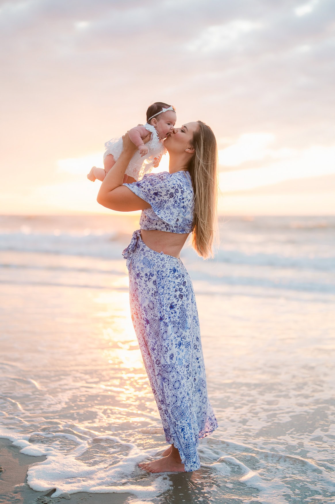 Beautiful family beach portrait at sunset capturing a mother holding her smiling baby girl on the shore with gentle ocean waves, perfect for family photography sessions.