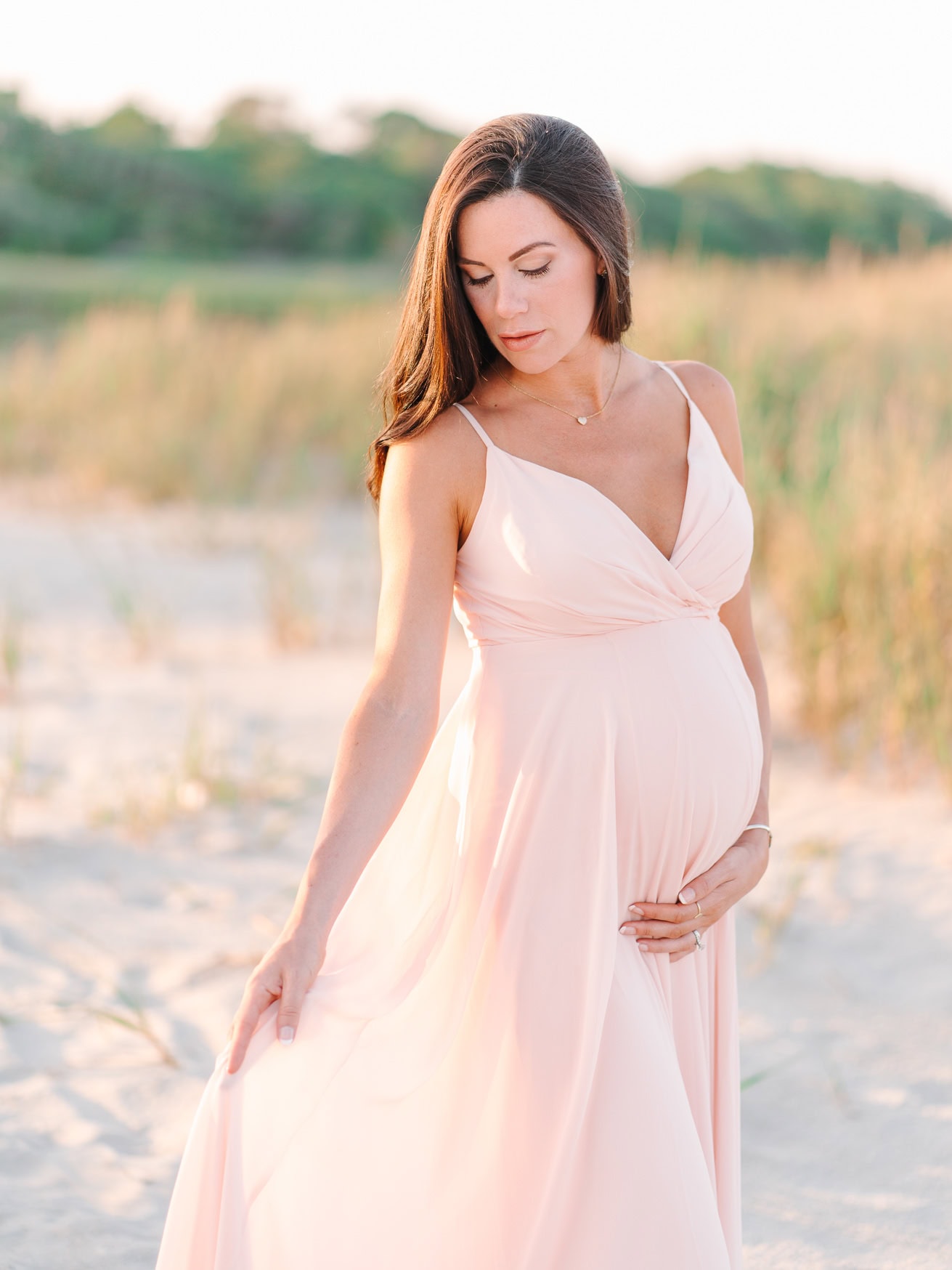 Elegant pregnant woman in a flowing blush pink maternity dress on the beach during sunset, capturing maternity photography, family beach portraits, and special moments with nature.