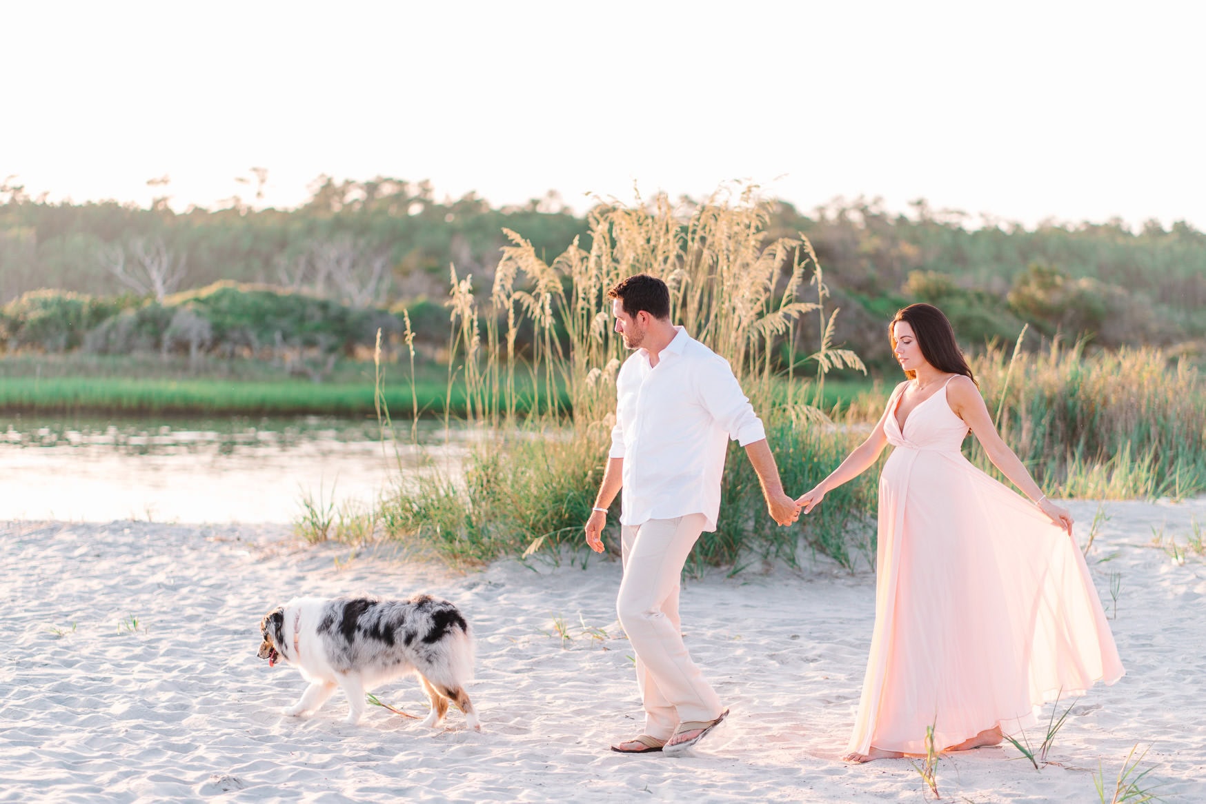 Beautiful couple holding hands walking on beach with dog during sunset, romantic coastal engagement or family beach portrait photography, soft pastel colors, natural light, outdoor setting.