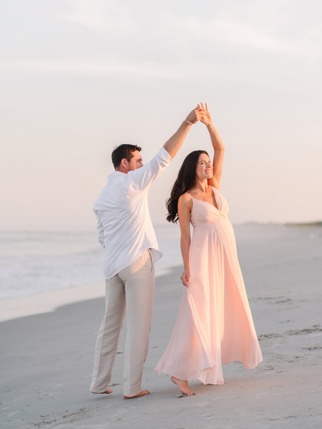 Romantic couple dancing on the beach at sunset, maternity photoshoot, elegant woman in a flowing pink dress, joyful moment by the sea, professional wedding and family beach portraits.