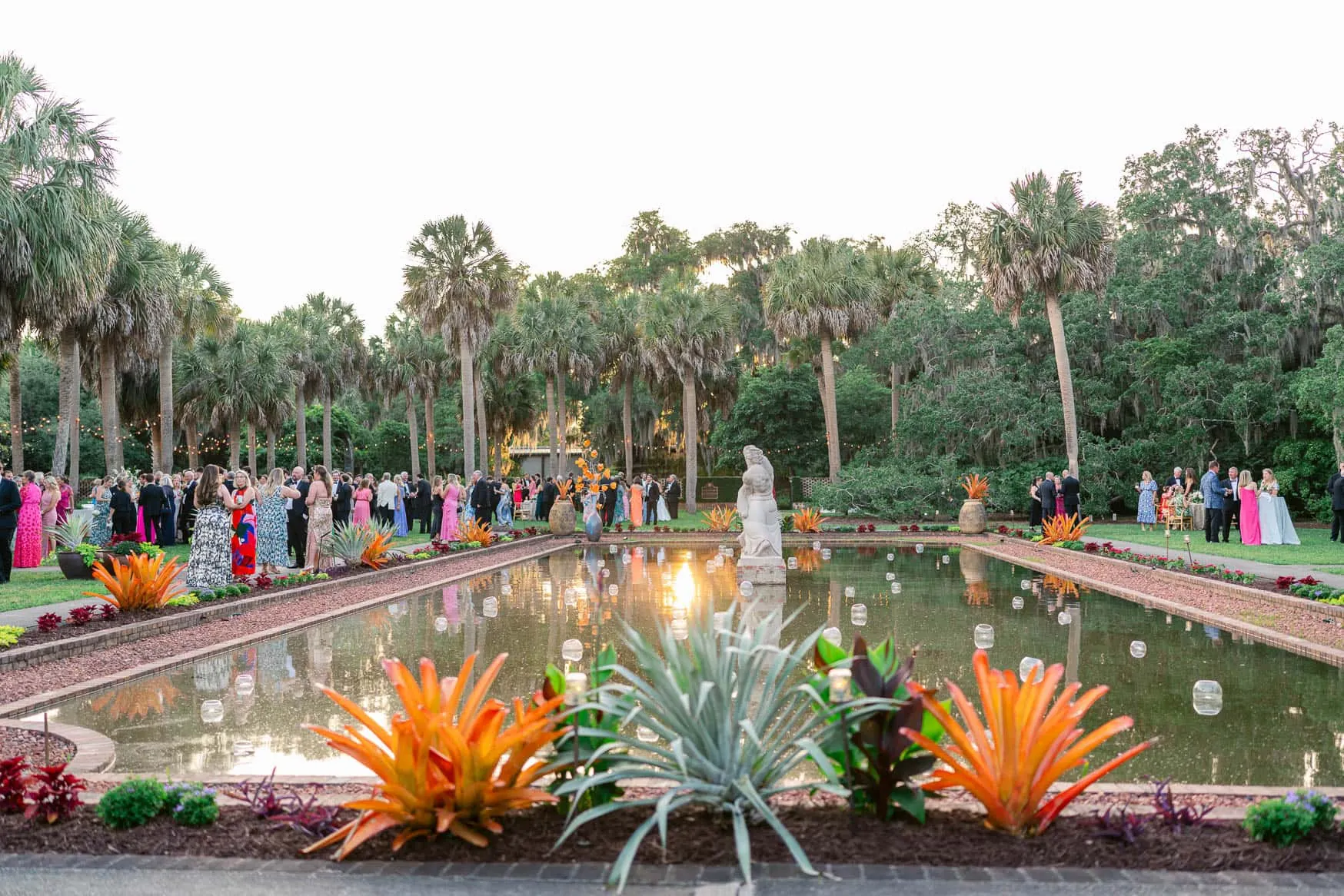 Colorful outdoor wedding reception by a fountain with tropical plants and tall palm trees at sunset, lively celebration with guests in formal attire, romantic and scenic event photography.