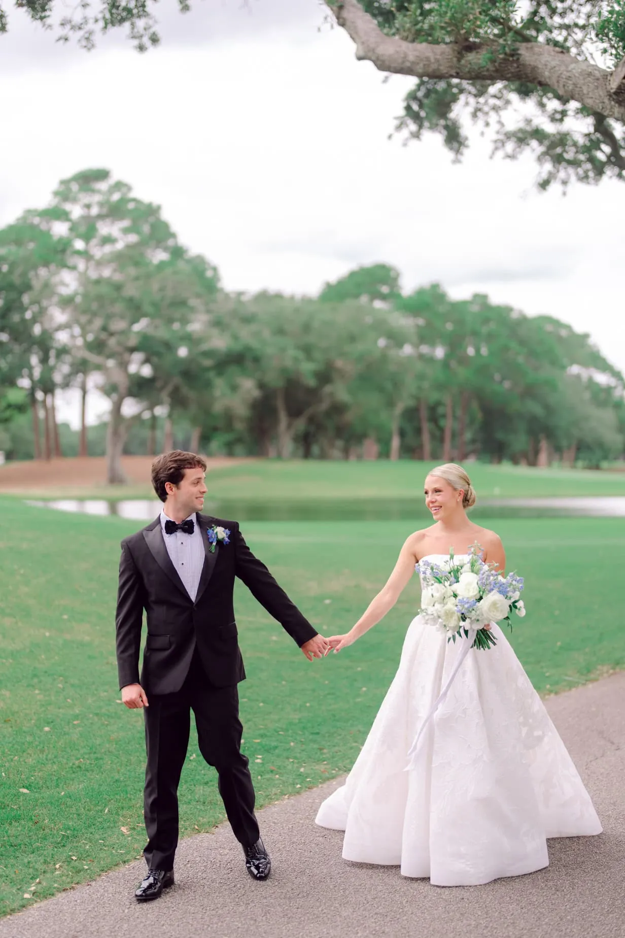 Elegant wedding portrait of a bride and groom holding hands on a lush green golf course with trees and water in the background.