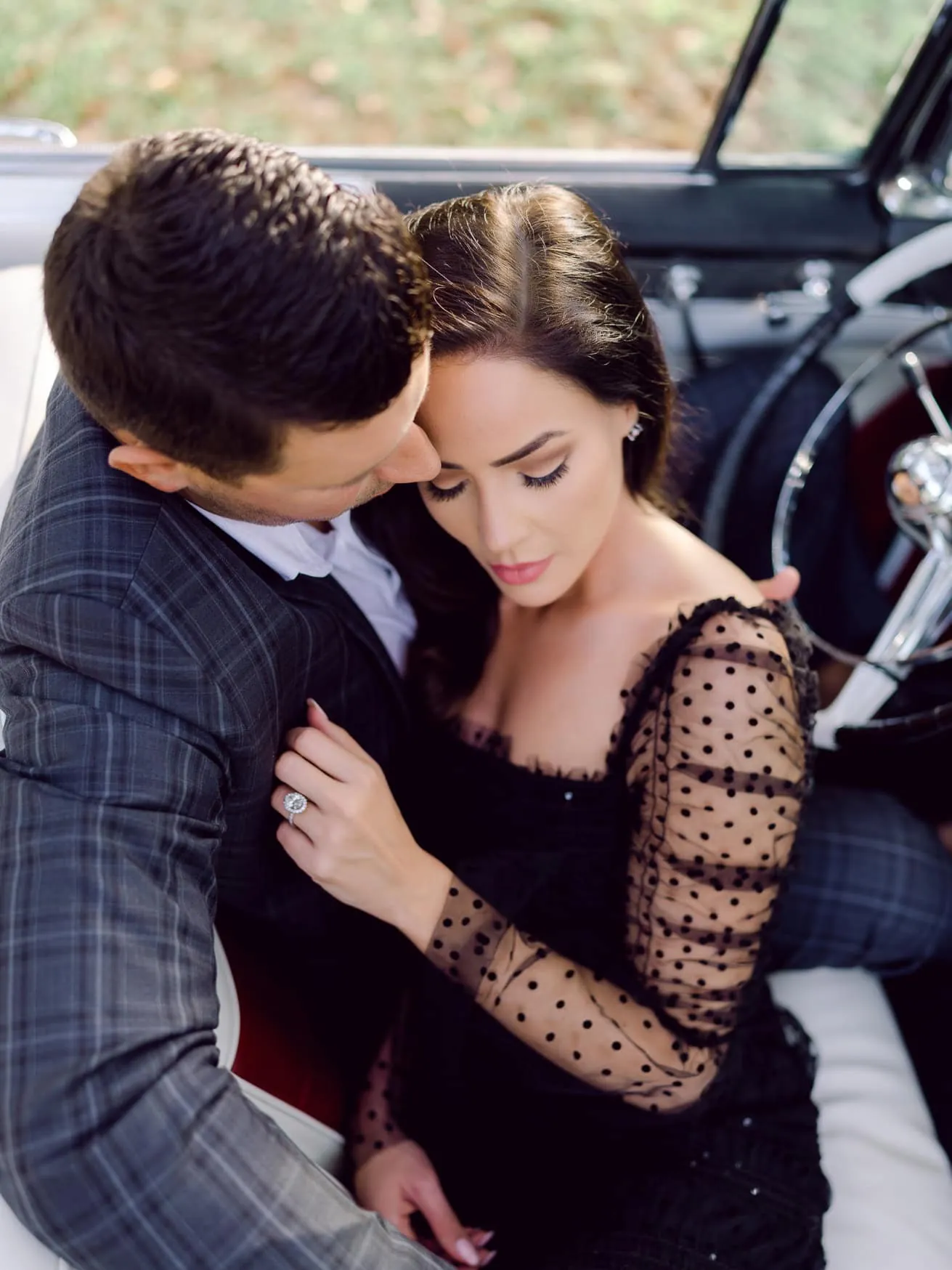 Romantic couple portrait in vintage car, woman wearing black polka dot dress, bride or engaged woman, soft natural light, intimate wedding or engagement photography, elegant and stylish.
